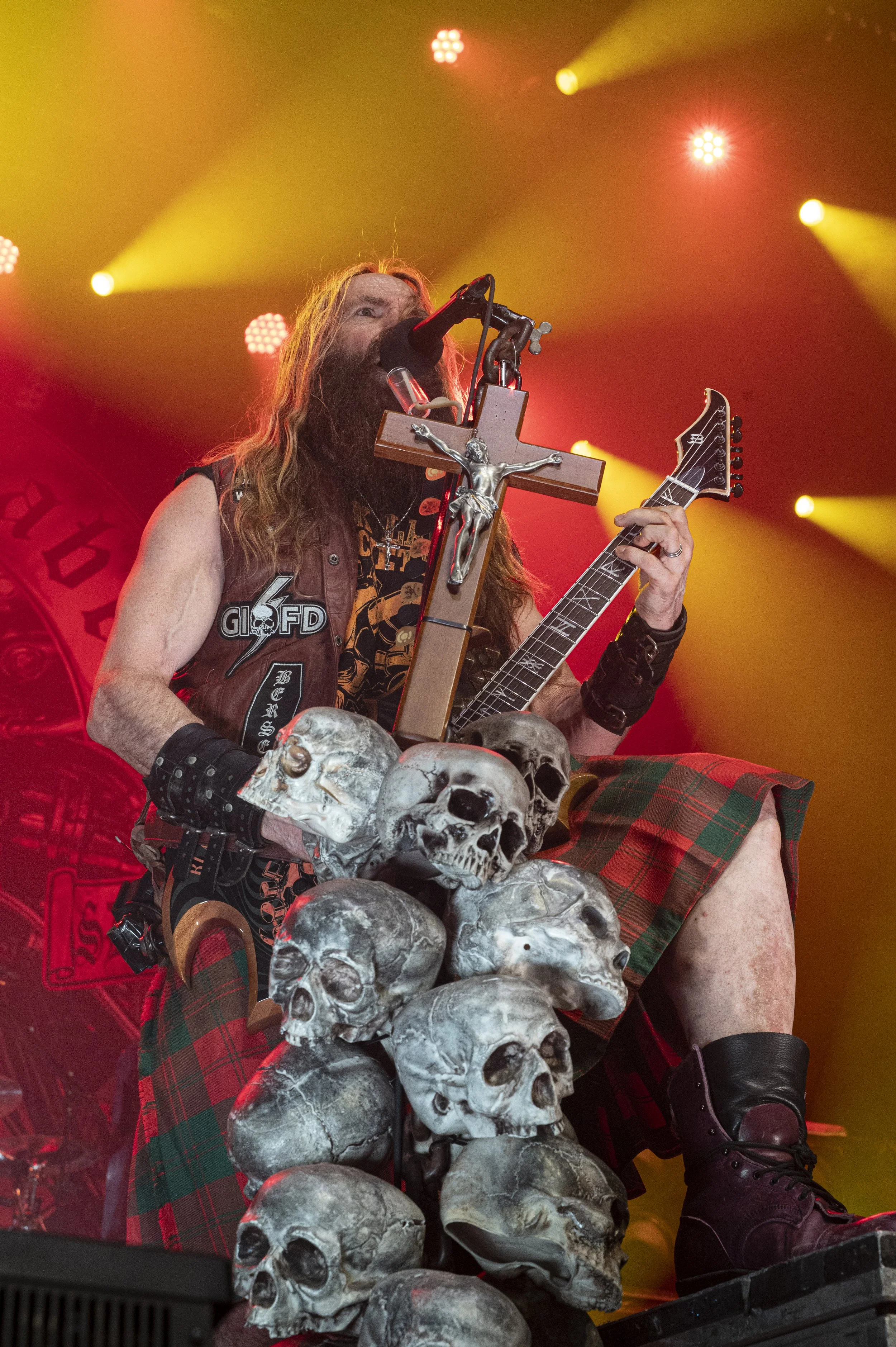 A musician performing on stage with a microphone, a cross with a Jesus figure, an electric guitar, and decorated with human skulls, dressed in a plaid kilt and black boots, under red and yellow stage lighting.