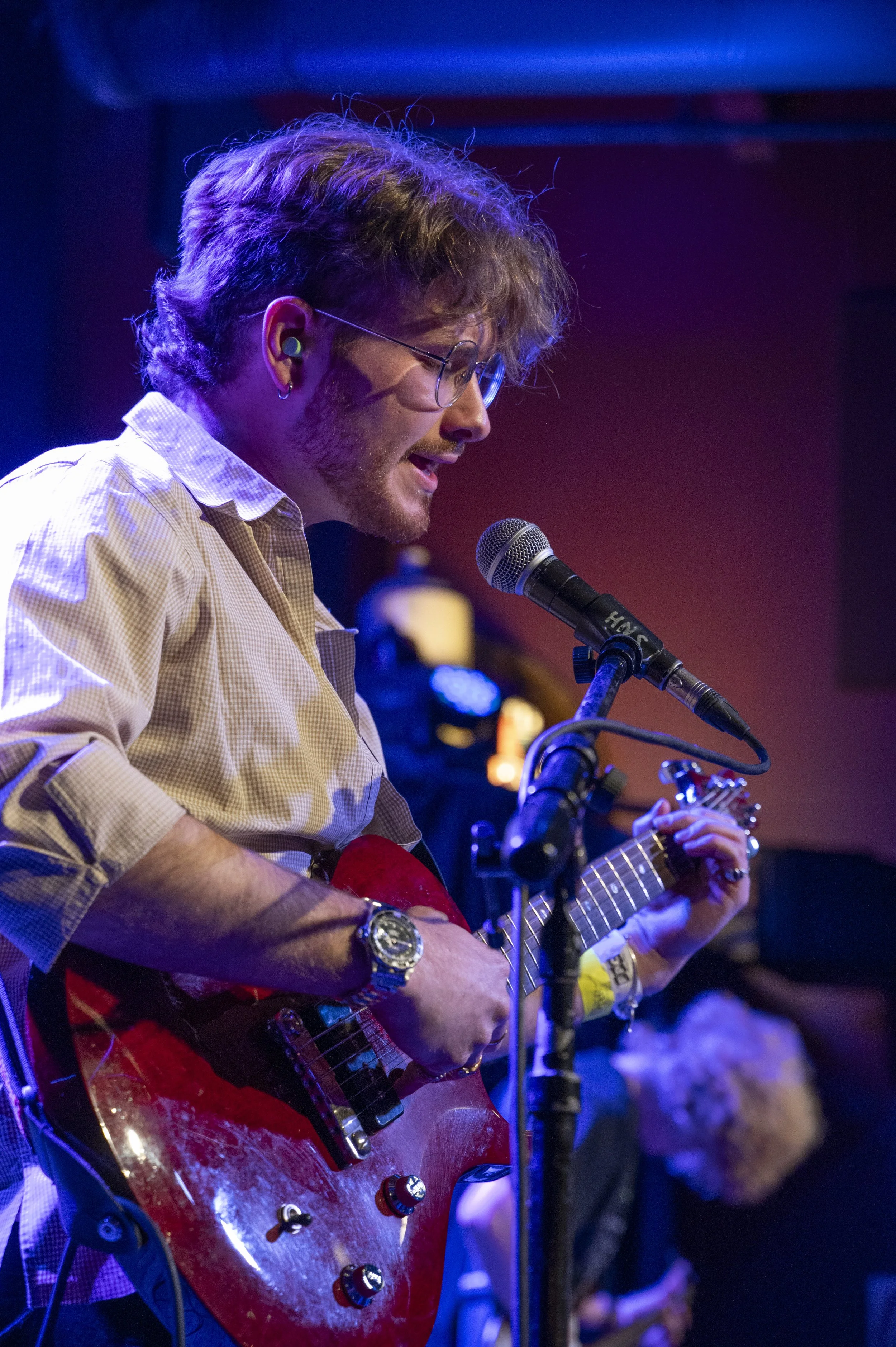 A man with glasses and curly hair performing on stage, playing an electric guitar and singing into a microphone, in a dimly lit setting with colorful stage lighting.