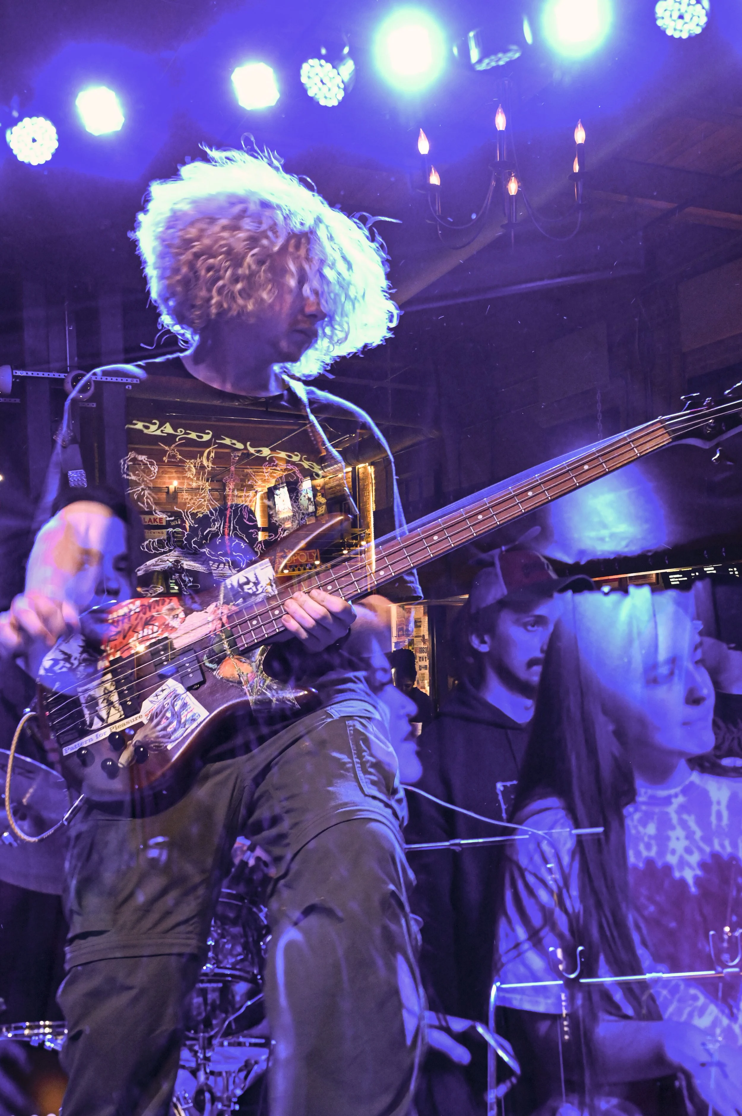 A man with curly blonde hair playing an electric bass guitar on stage under blue stage lights, with audience members visible in the background.