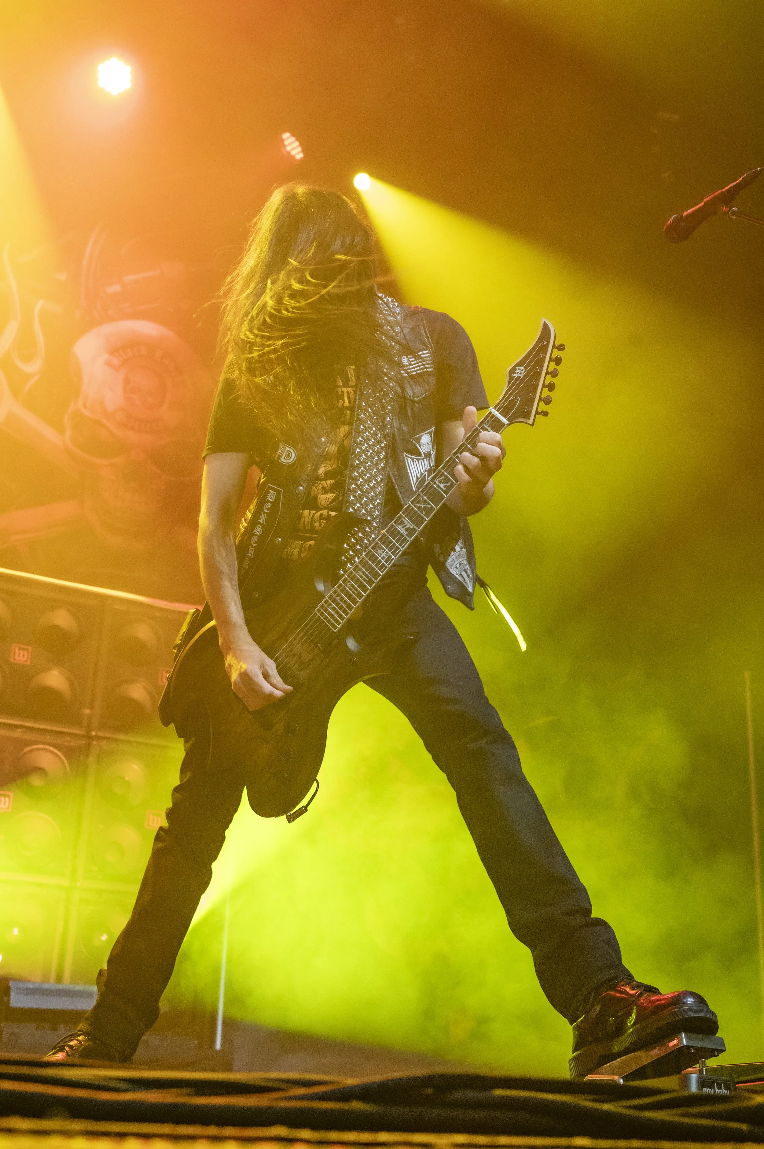 A guitarist performs on stage with yellow and orange lighting, wearing a black vest with patches and a studded scarf, and playing a dark electric guitar.