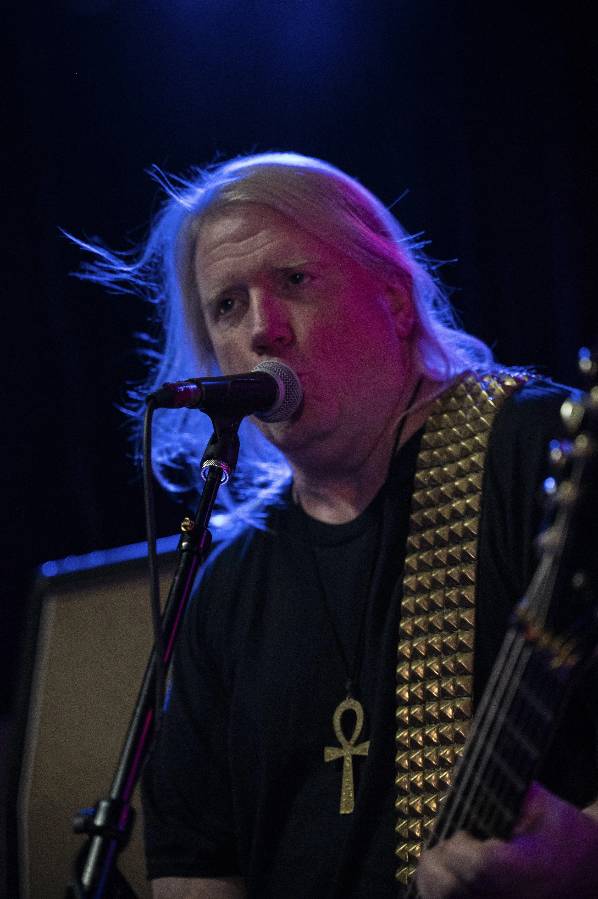 A male musician with long, platinum blonde hair singing into a microphone. He is wearing a black shirt, a necklace with an ankh pendant, and a guitar strap with gold spikes. The background is dark, with stage lighting creating a colorful glow on his 