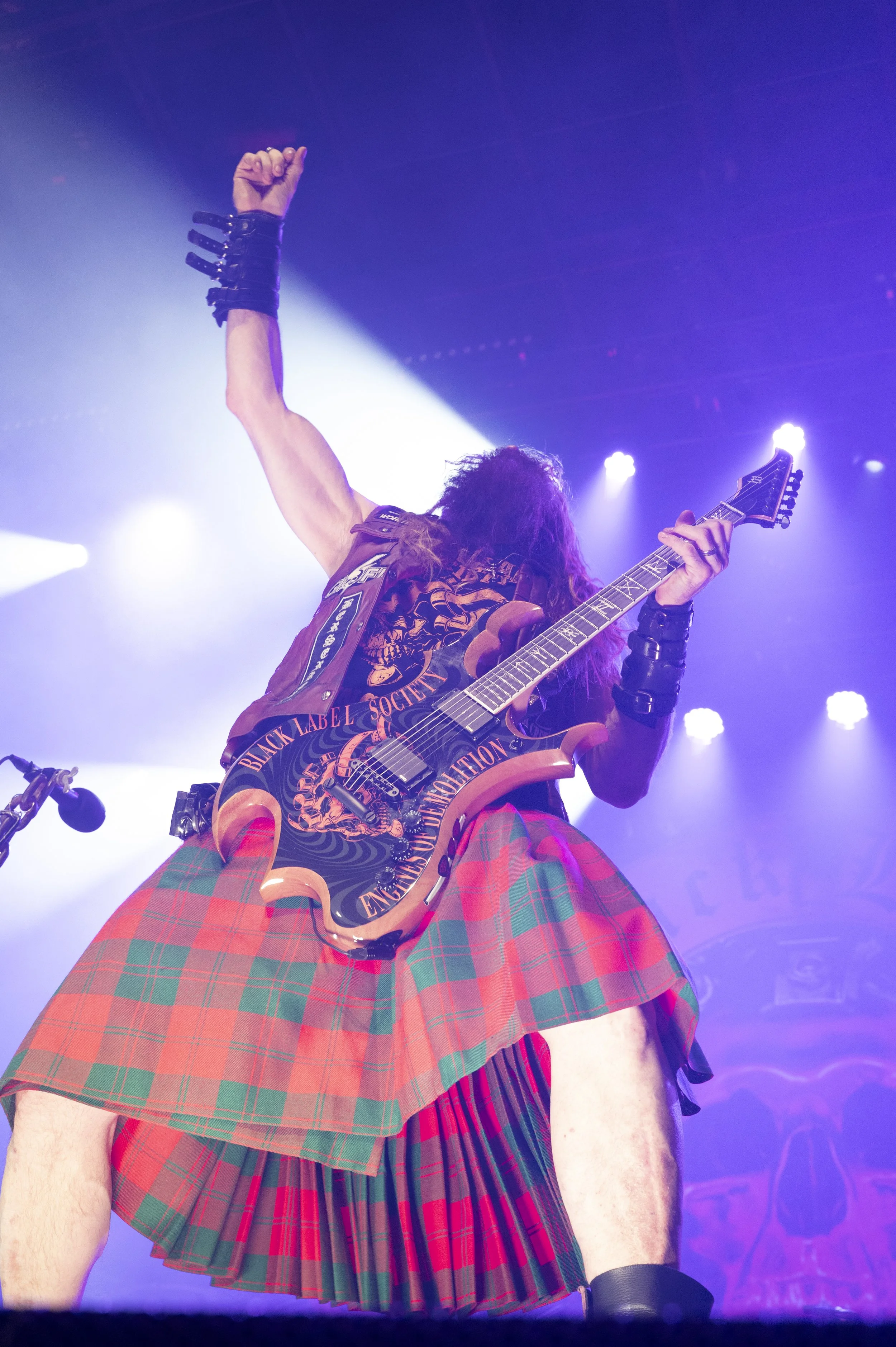 A guitarist on stage, wearing a kilt, playing an electric guitar with a design reading 'Black Label Society,' with stage lights shining down.