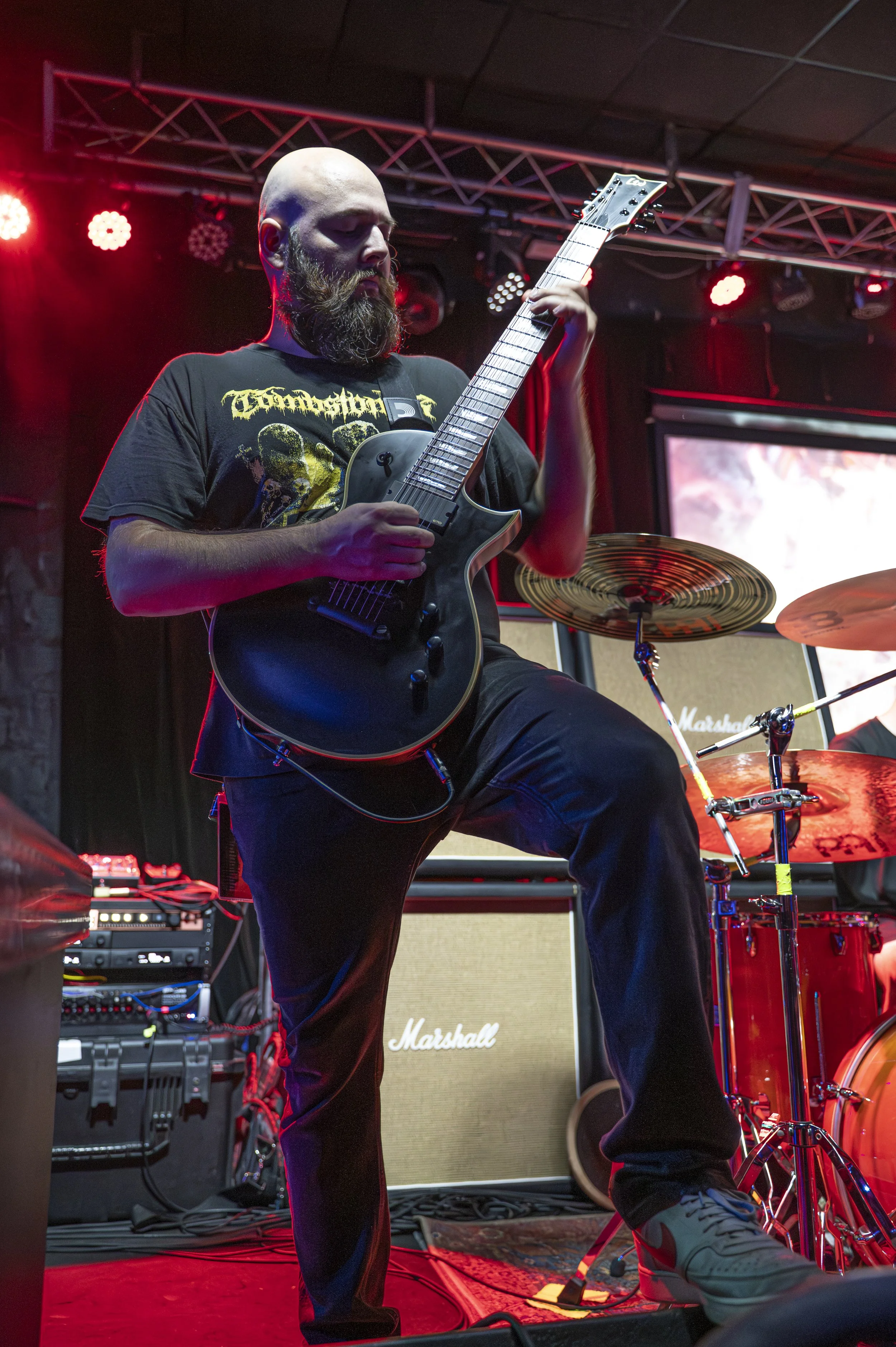 A bearded man playing an electric guitar on stage with red stage lights, a Marshall amplifier, and drum set behind him.