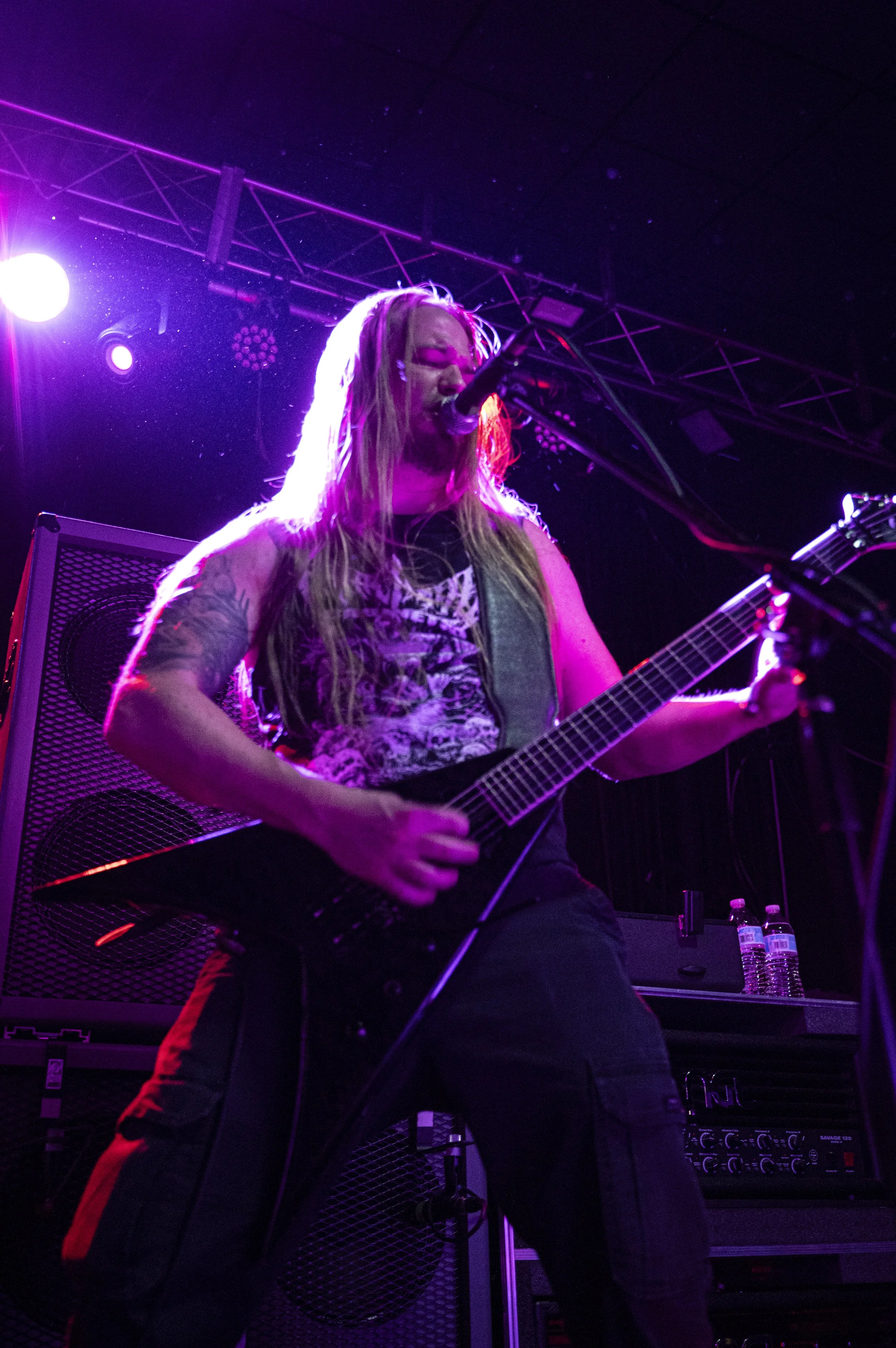 A musician with long blond hair and tattoos, wearing a graphic t-shirt and black pants, performing with an electric guitar on stage under purple lighting.