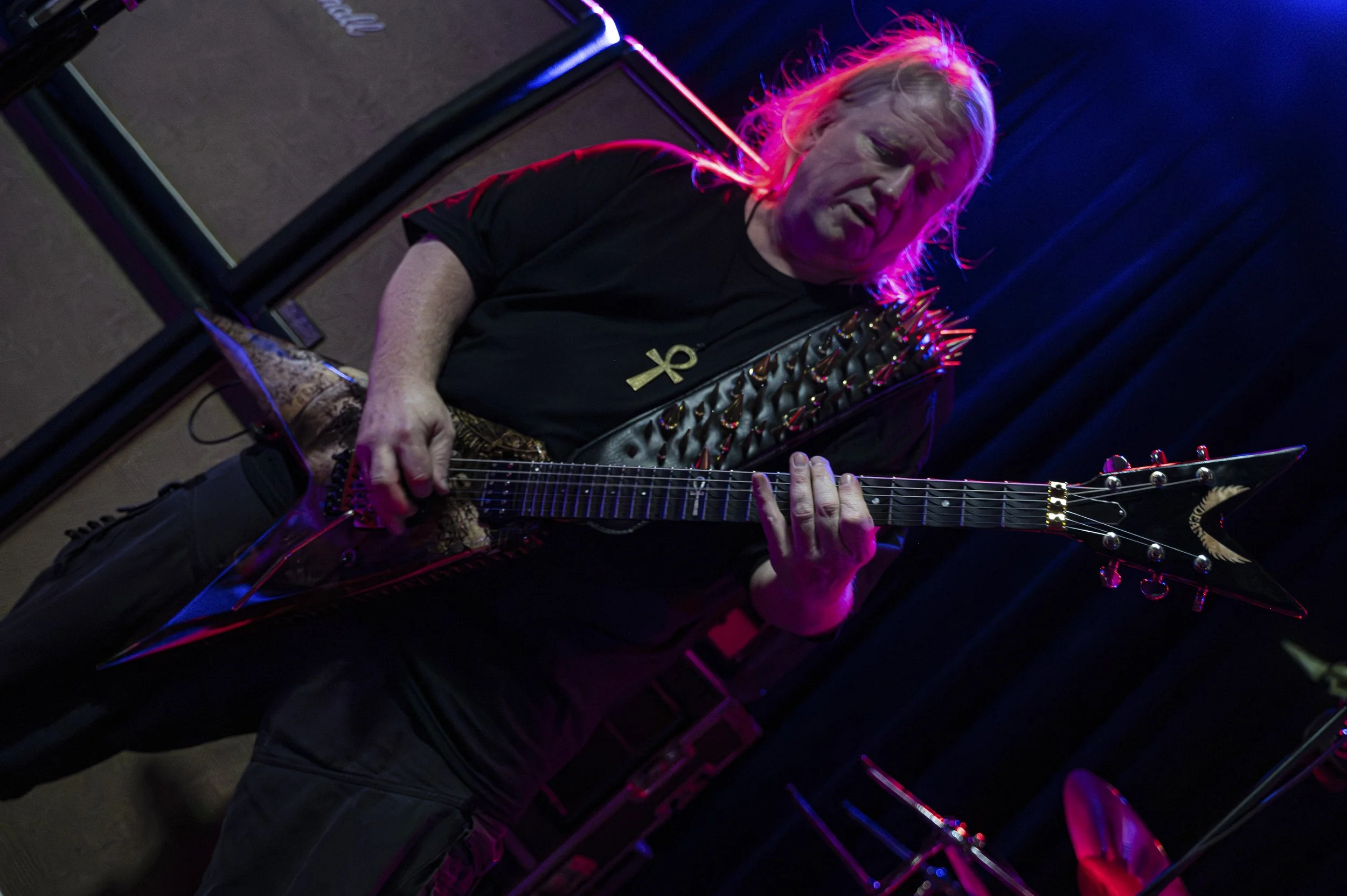 A man with long blond hair playing a black electric guitar with a spiked design on stage, illuminated by bright pink and purple lights, with black curtains in the background.
