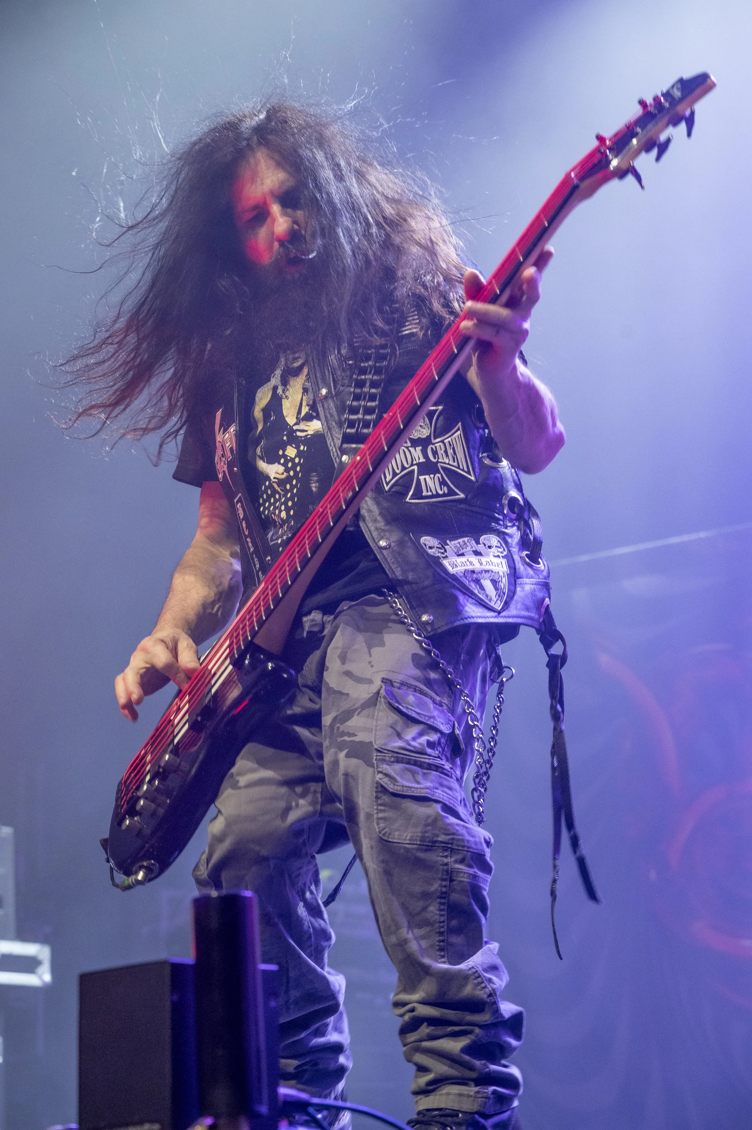A male guitarist with long, messy hair, wearing a black leather vest and cargo pants, playing a red electric guitar on stage with purple lighting.