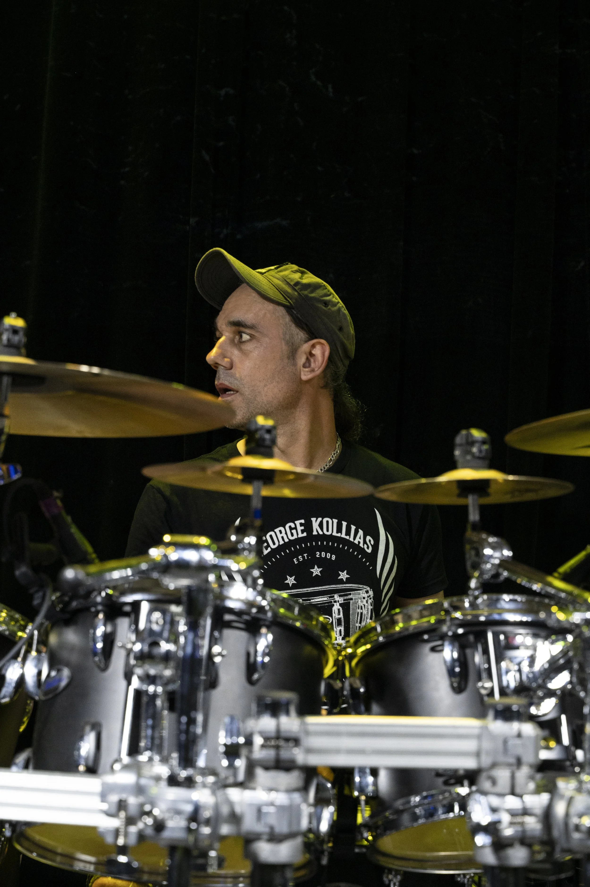 A man with a dark cap and a black T-shirt sitting behind a drum set, looking to his left against a dark background.