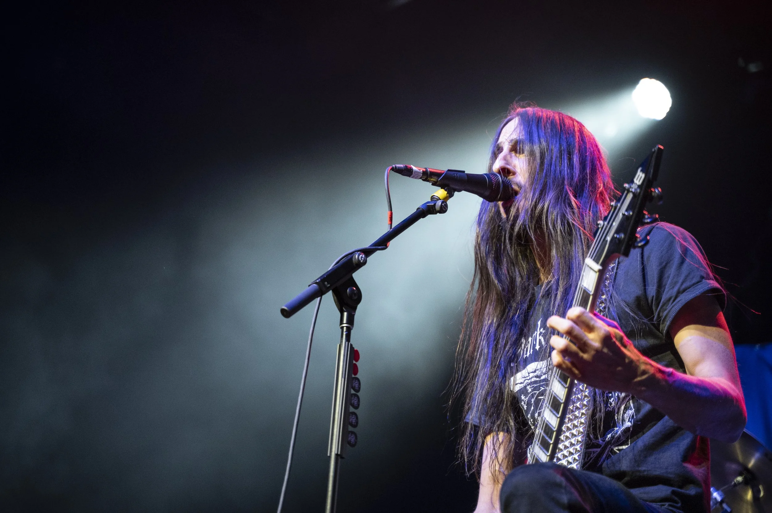 A long-haired musician singing into a microphone while playing an electric guitar on stage with bright stage lights.
