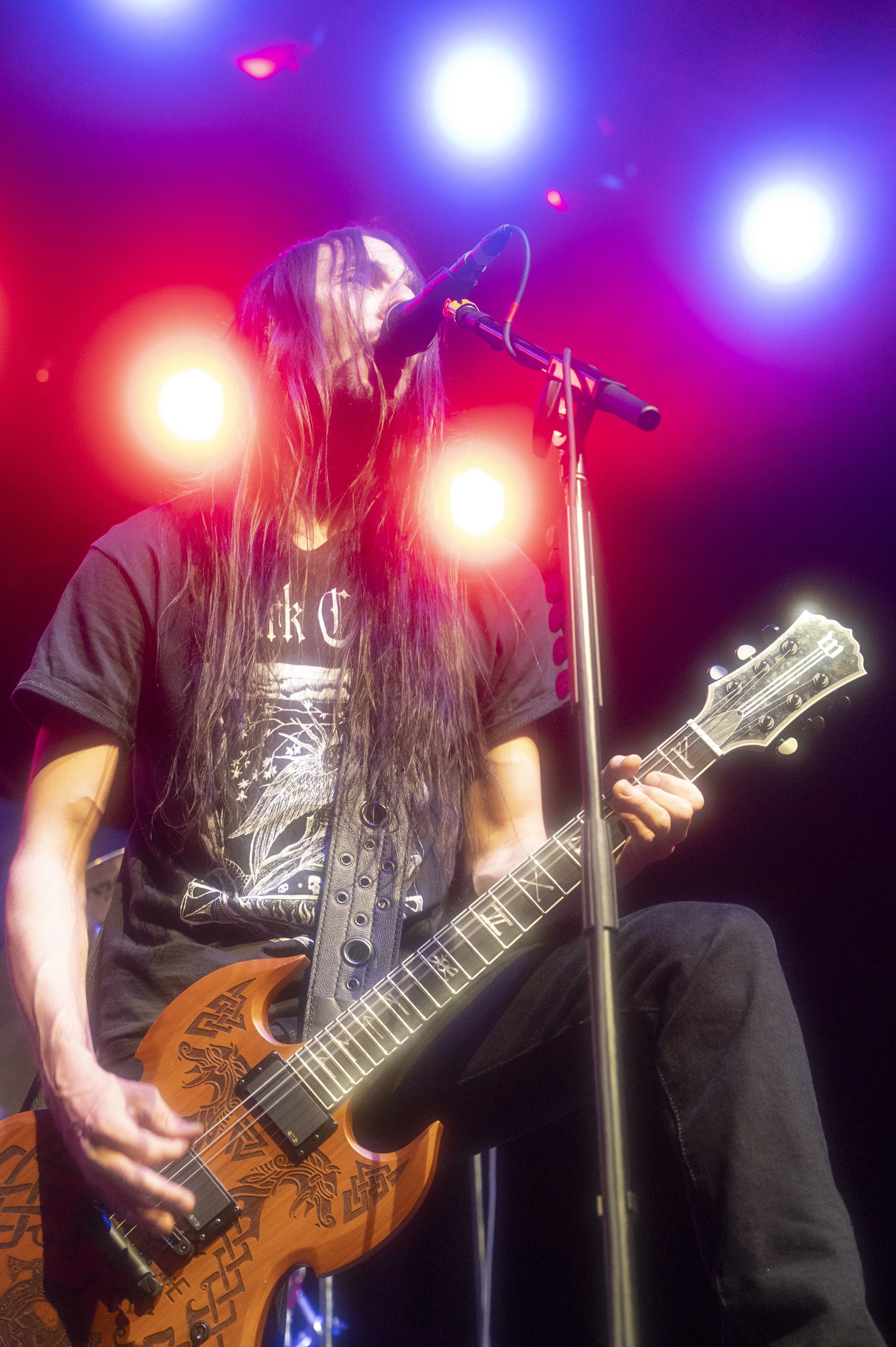 A rock musician with long dark hair playing an electric guitar and singing into a microphone on stage under colorful lights.