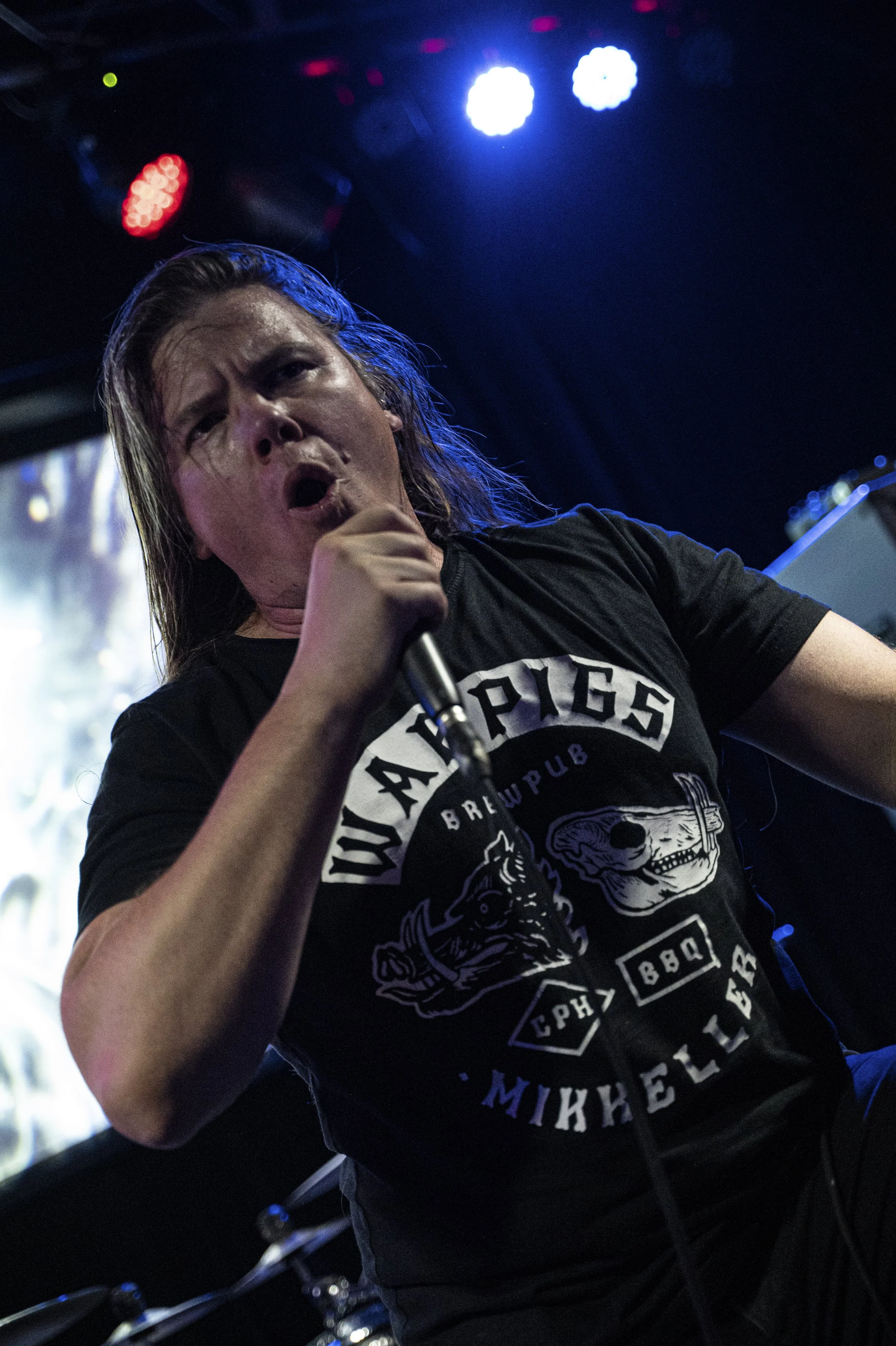 Performer singing into a microphone on stage, wearing a black T-shirt with a graphic design and the words 'WARRIORS Brewpub' and 'Michelle' in white and blue letters, with colorful stage lights overhead.