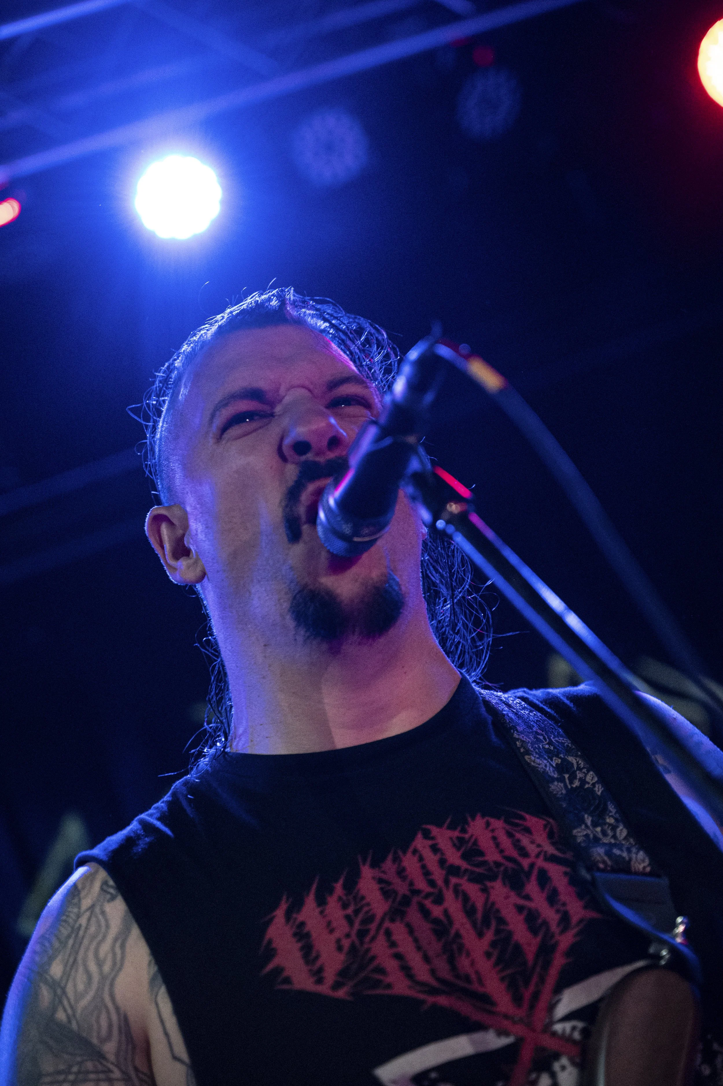 A male musician singing into a microphone on stage, illuminated by colorful stage lights, wearing a sleeveless shirt with a red graphic design and tattoos on his arm.