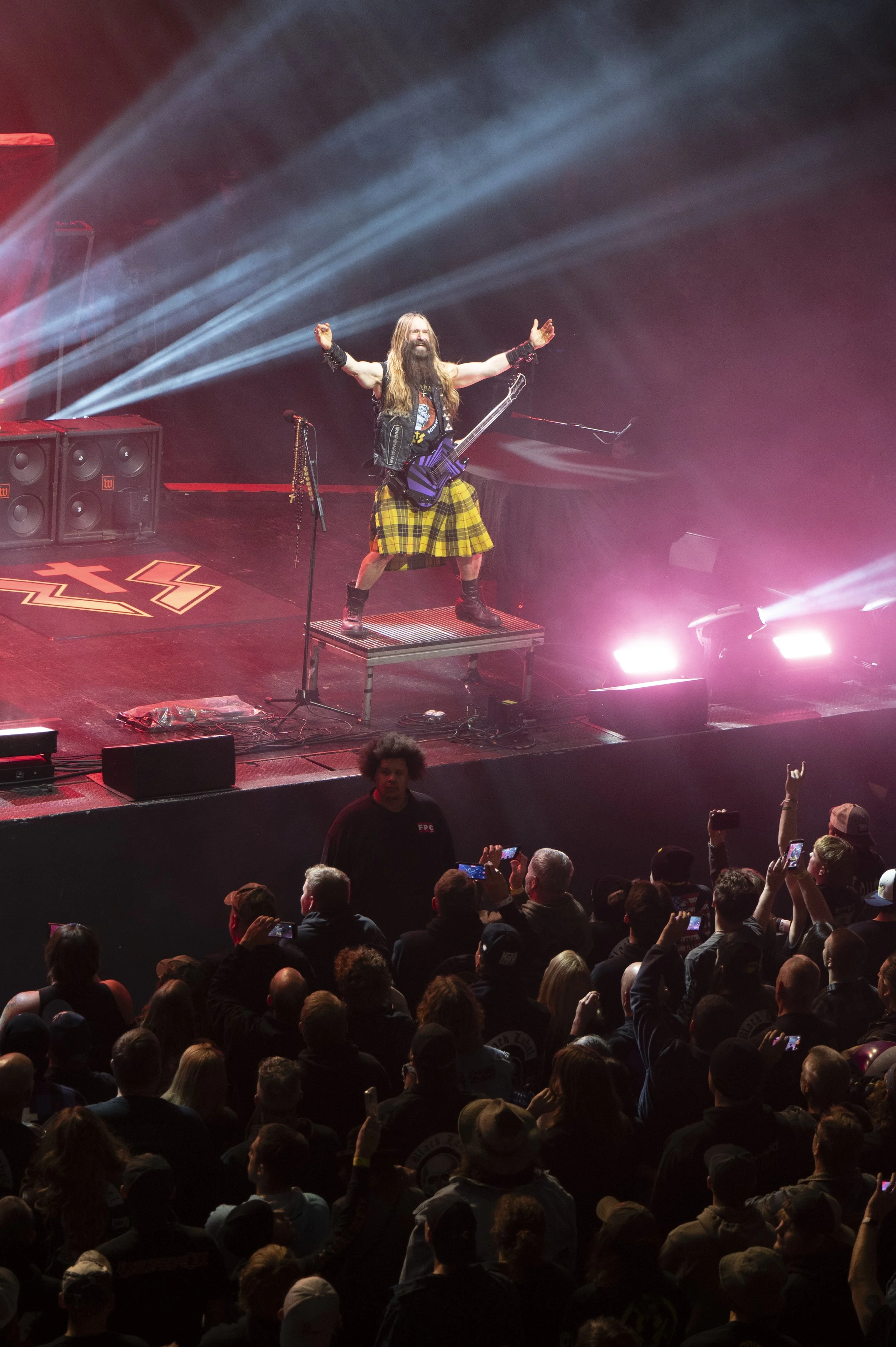 A rock musician with long hair, beard, and a kilt, standing on a raised platform on a stage, with arms raised, performing for a large audience. The stage is lit with colorful lights and the crowd is taking photos.