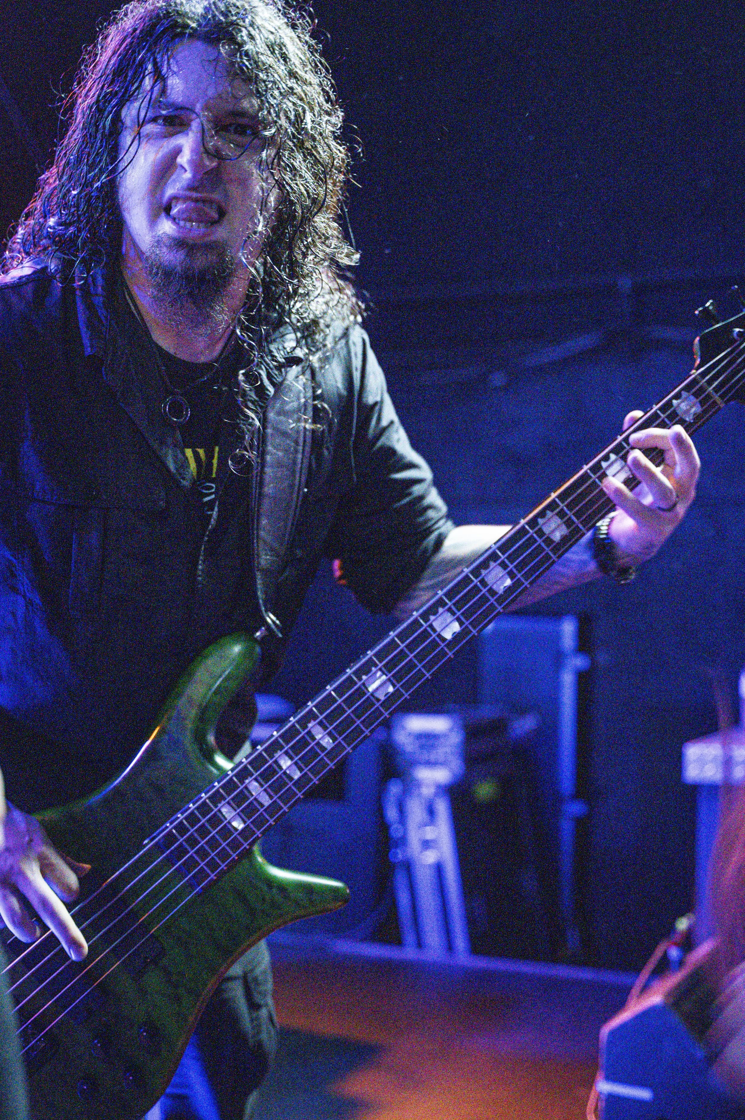 A man with long curly hair and a goatee, wearing a black leather jacket, is playing a green bass guitar on stage with a dark background and blue lighting.
