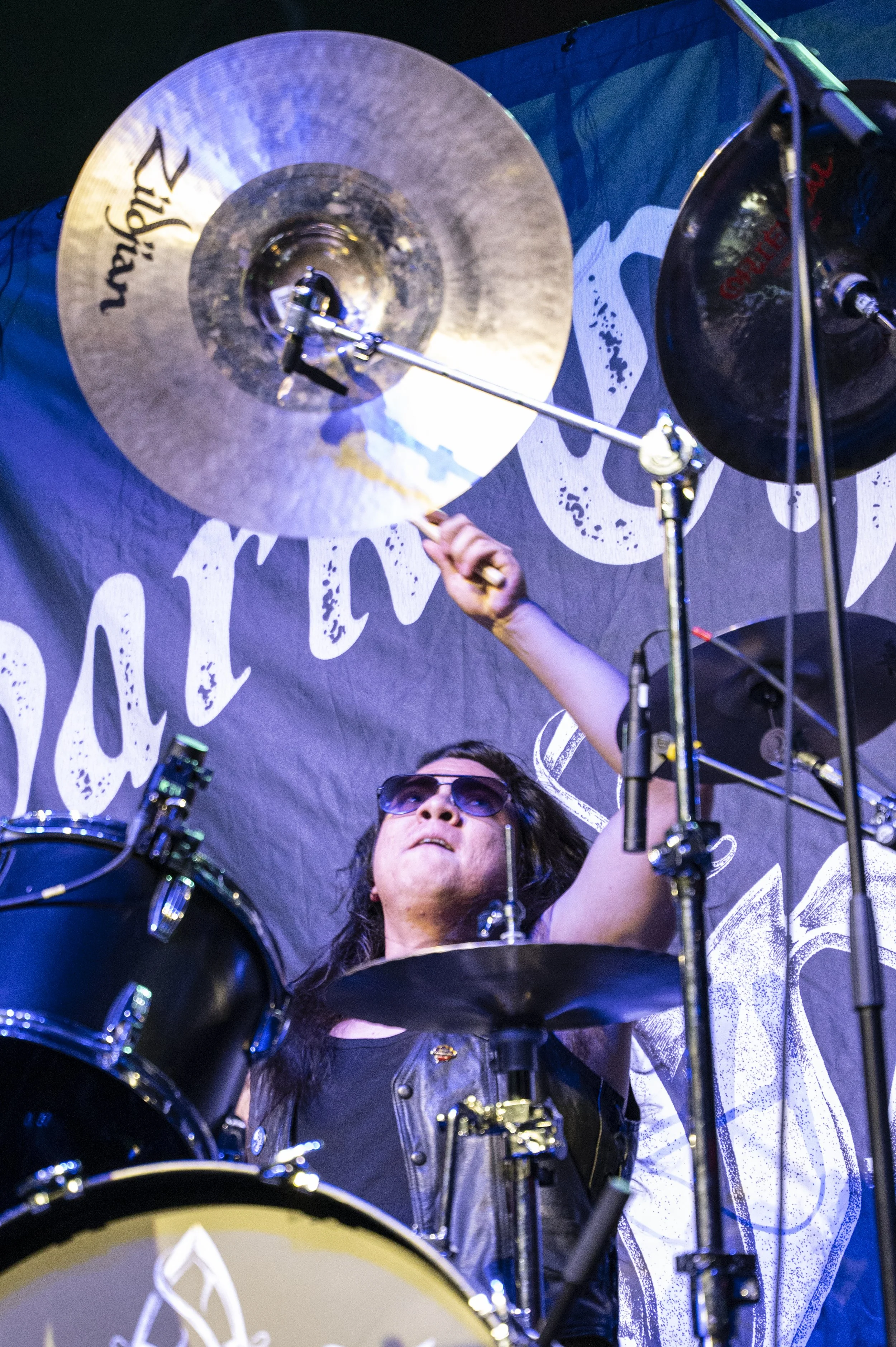 A drummer wearing sunglasses and a black vest playing a drum set on stage, with a black banner behind him featuring white text and design.