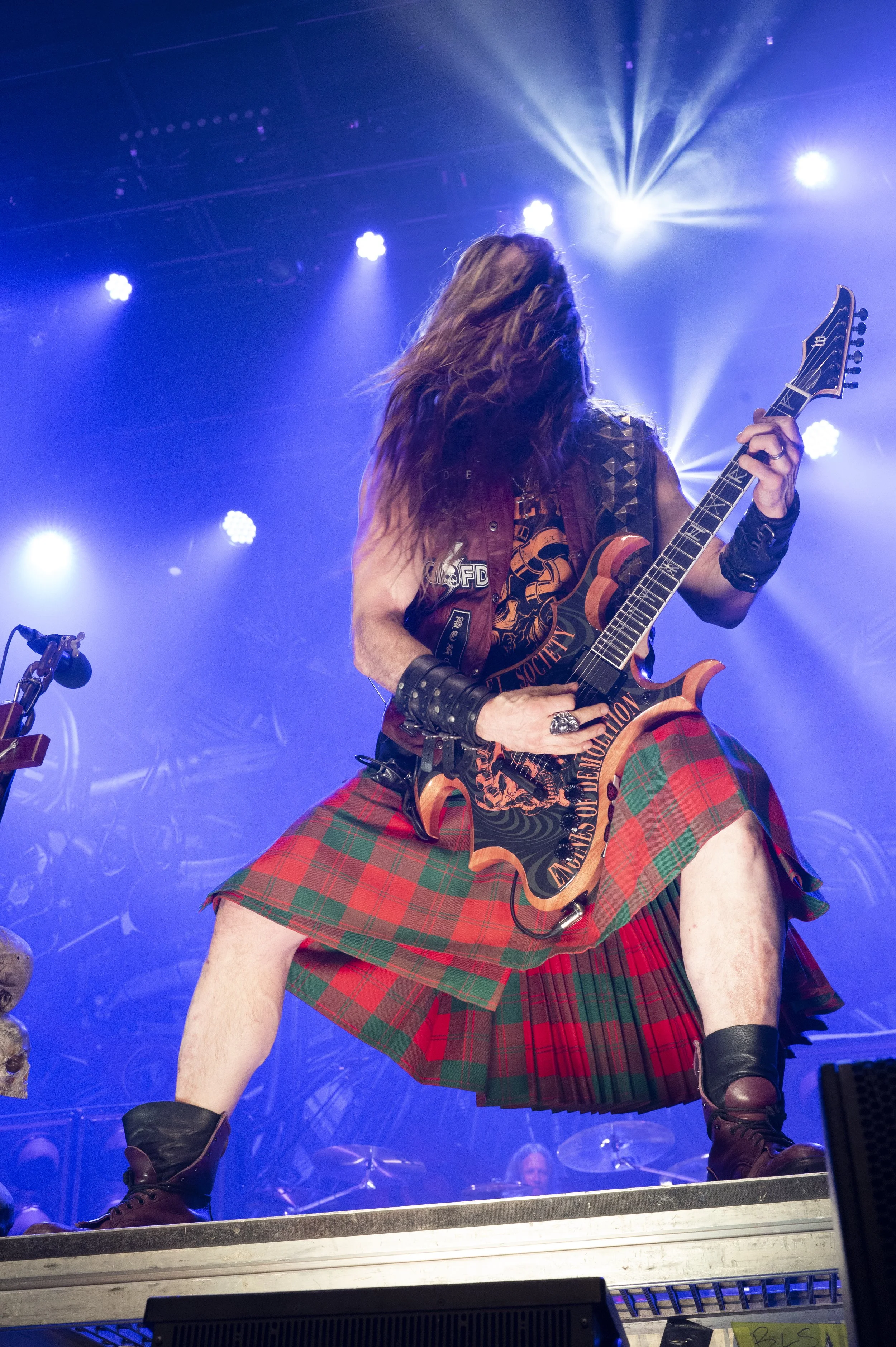 Heavy metal guitarist wearing a tartan kilt and leather accessories playing on stage with dramatic lighting.