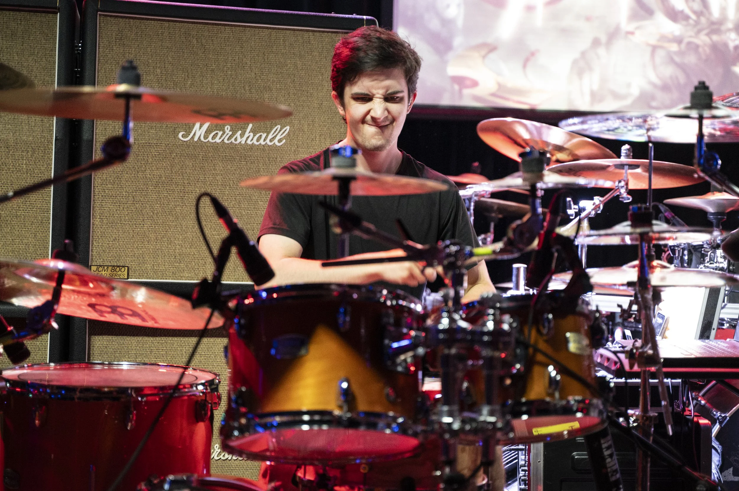 A young man with dark hair playing a drum set on stage, with a large Marshall amplifier behind him, surrounded by drums and cymbals.