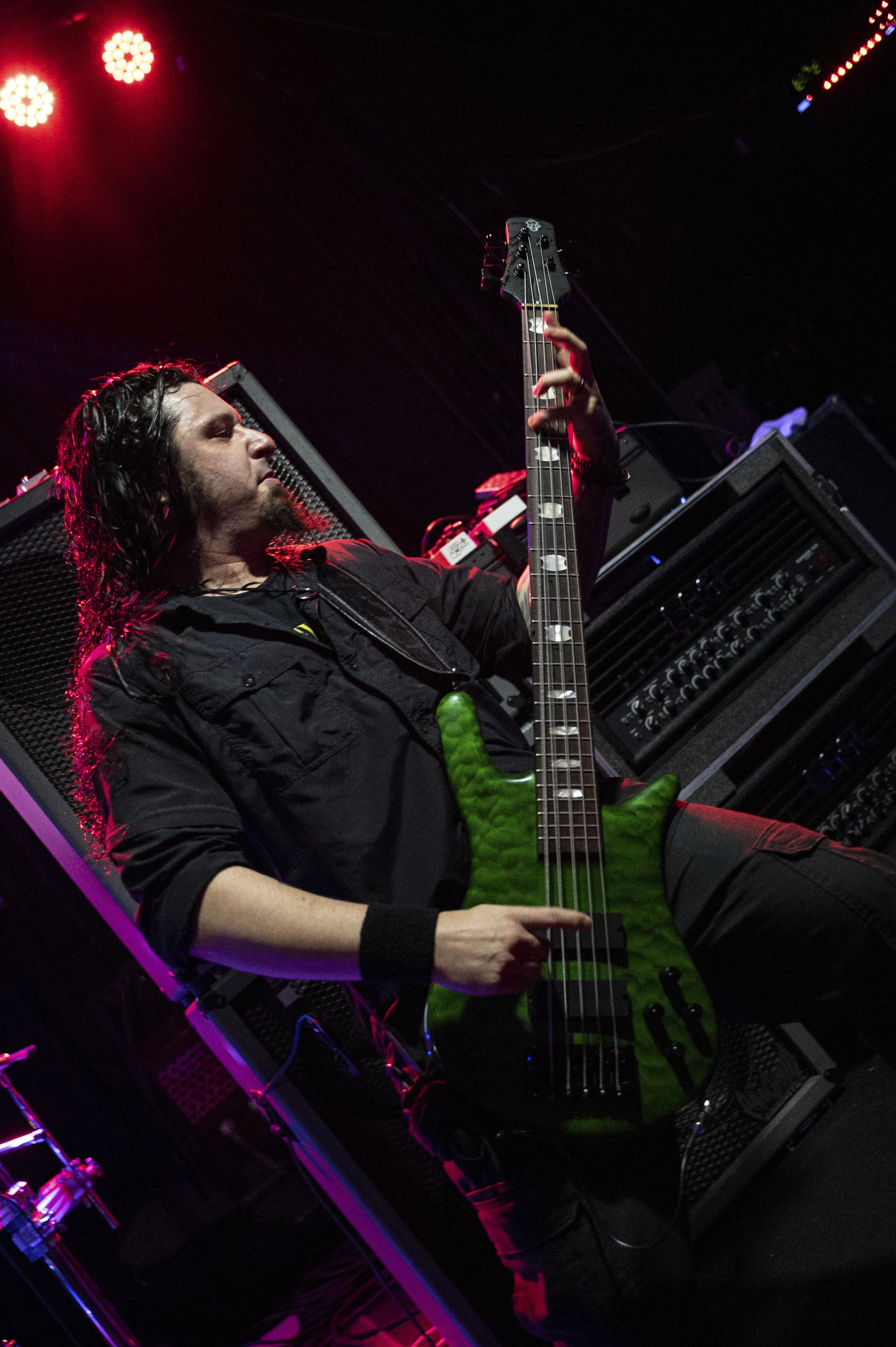 A musician with long dark hair playing a green electric bass guitar on stage under red and purple lights.