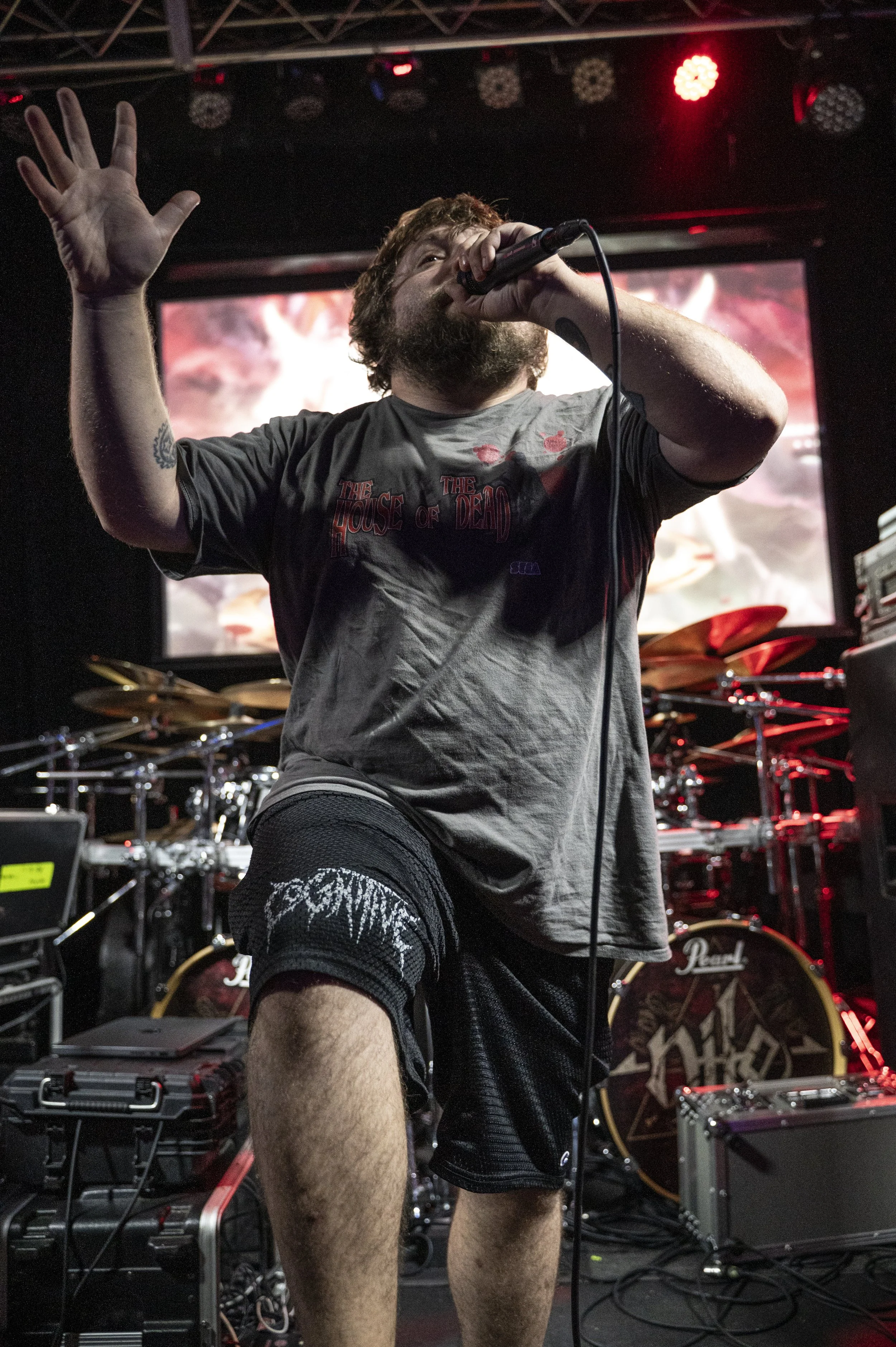 Male vocalist singing into a microphone on stage during a concert with drums and electronic equipment behind him.