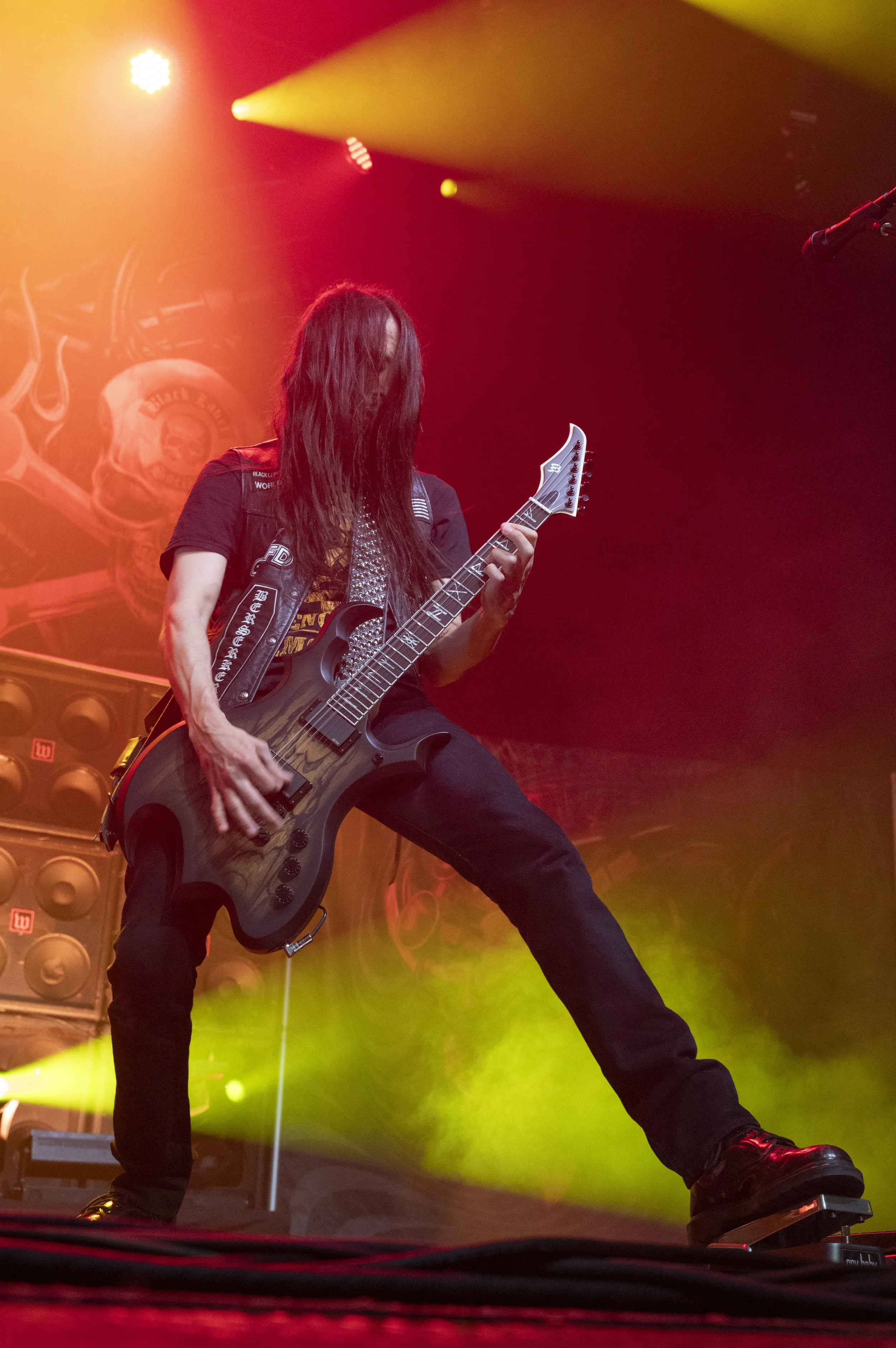 A long-haired guitarist performing on stage with colorful stage lighting and large speaker stacks in the background.