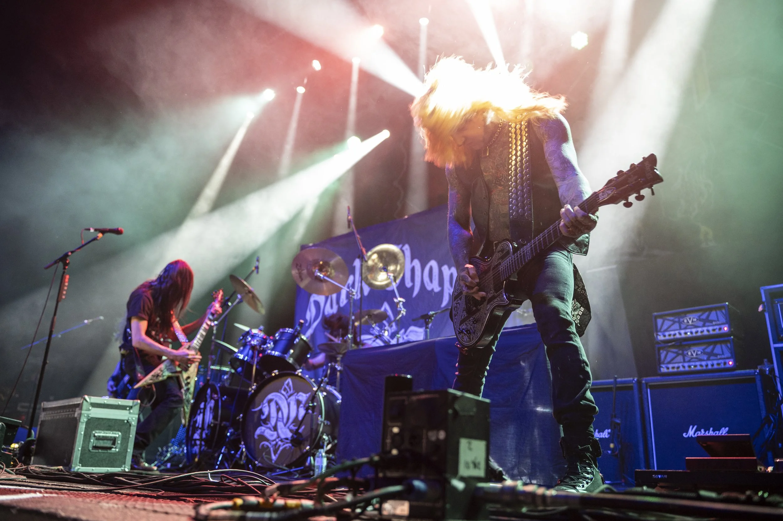 A rock band performing live on stage with colorful lighting, including two guitarists and a drummer, and a backdrop with the words 'Slash Harm'.