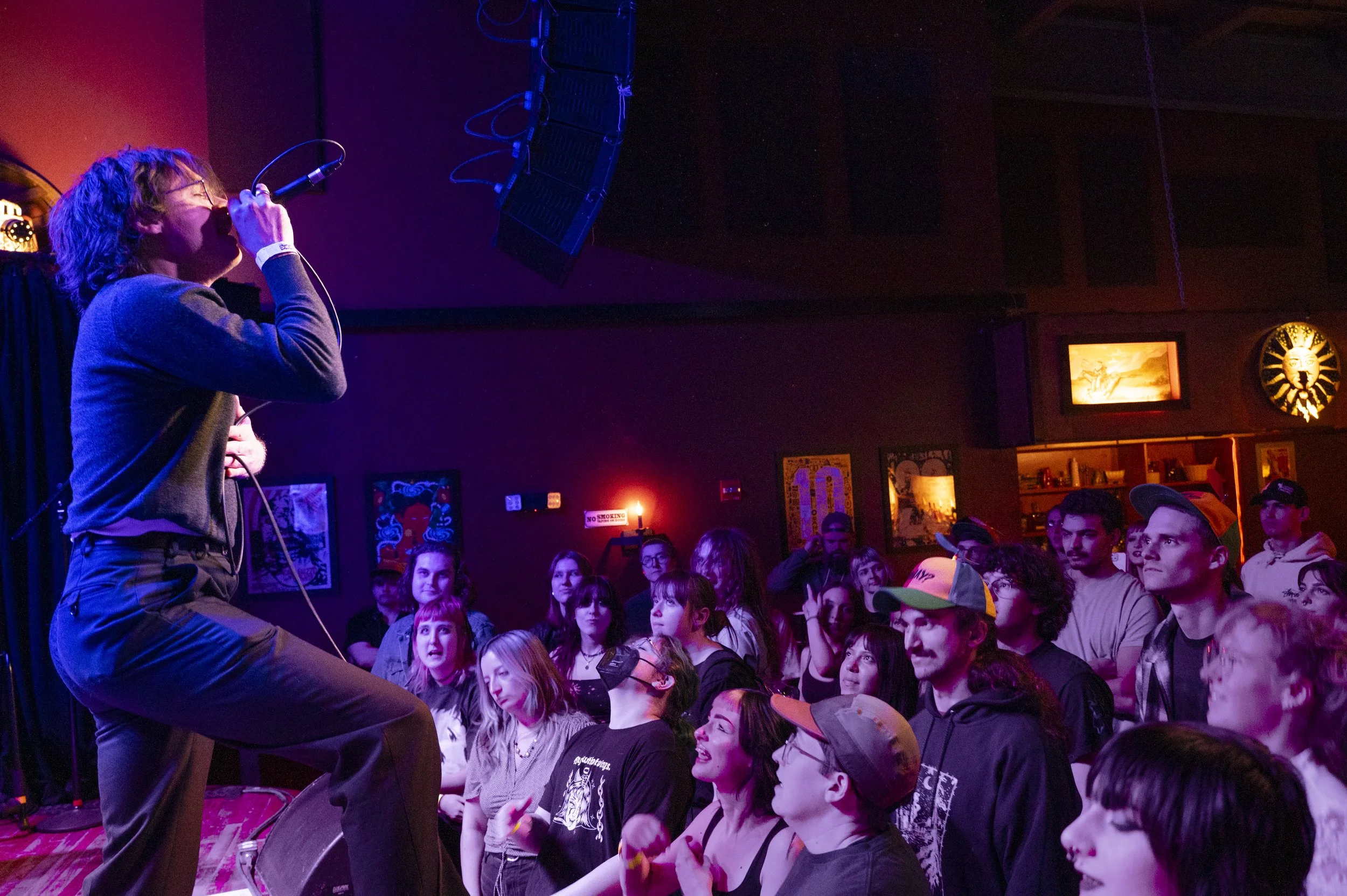 A musician singing into a microphone on stage at a concert or performance event, with an audience watching attentively.