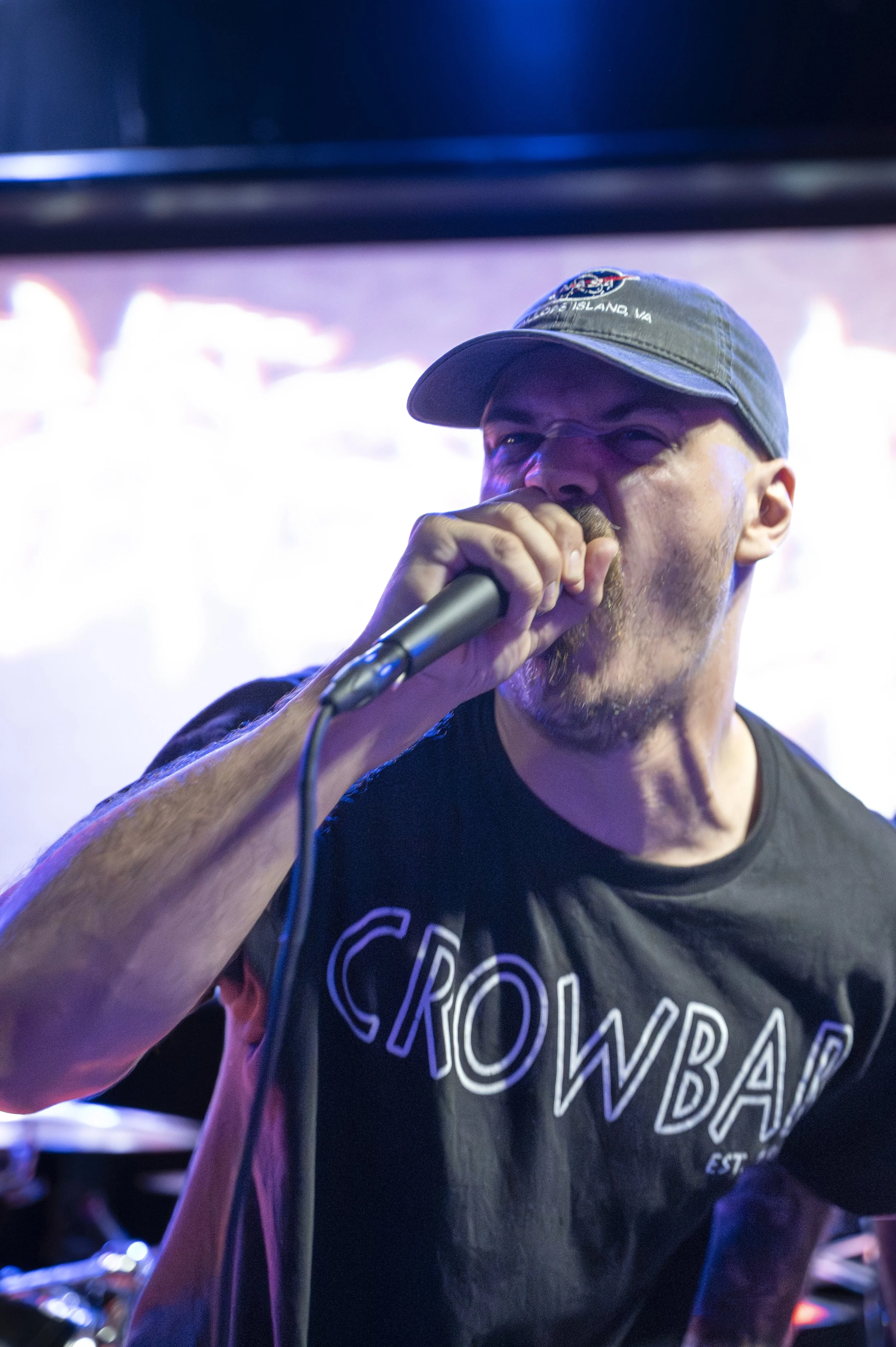 Man singing into a microphone at a live event, wearing a black T-shirt with the word 'CROWD