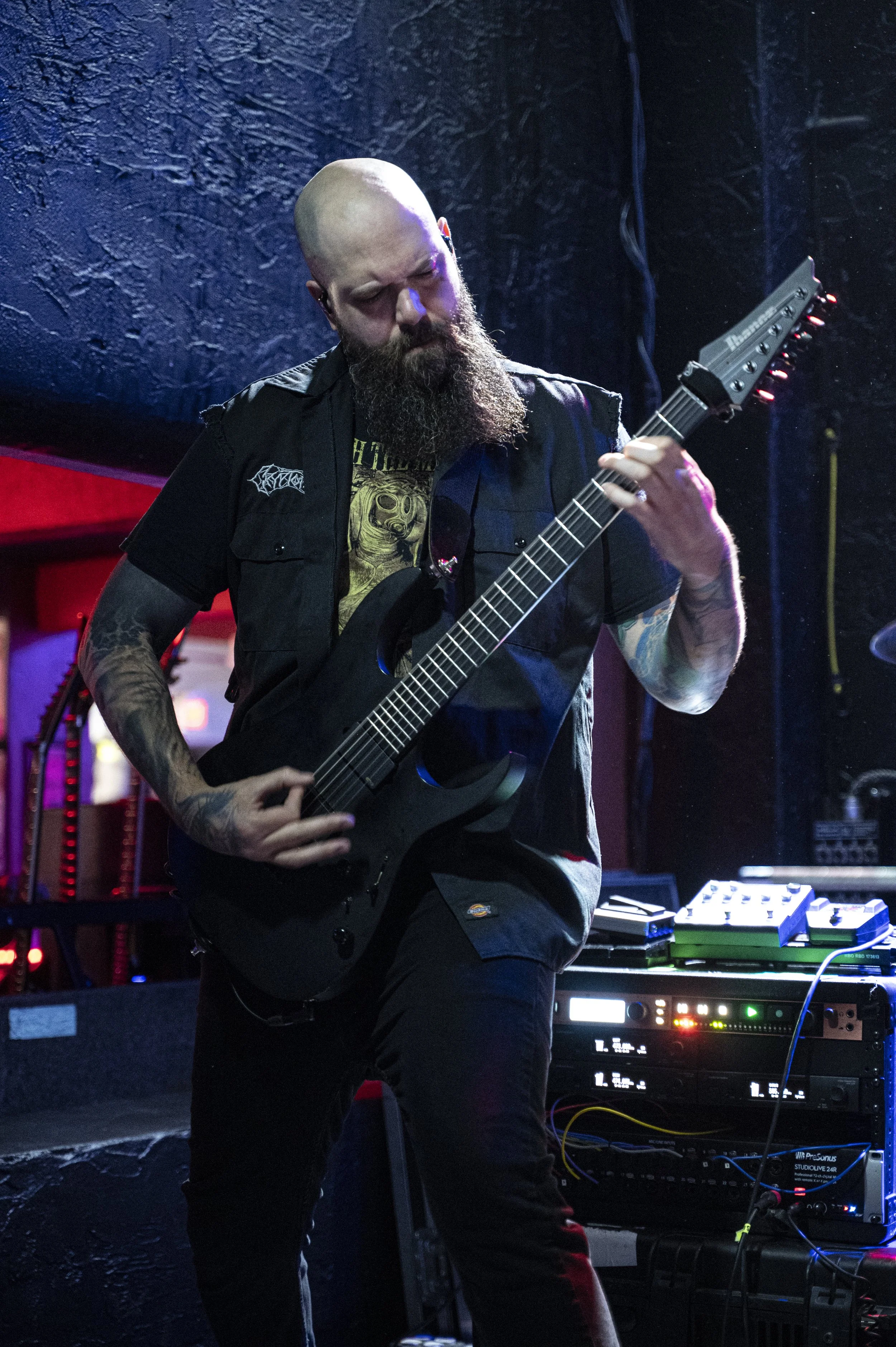 A bearded man with a shaved head playing an electric guitar on stage with music equipment in the background.