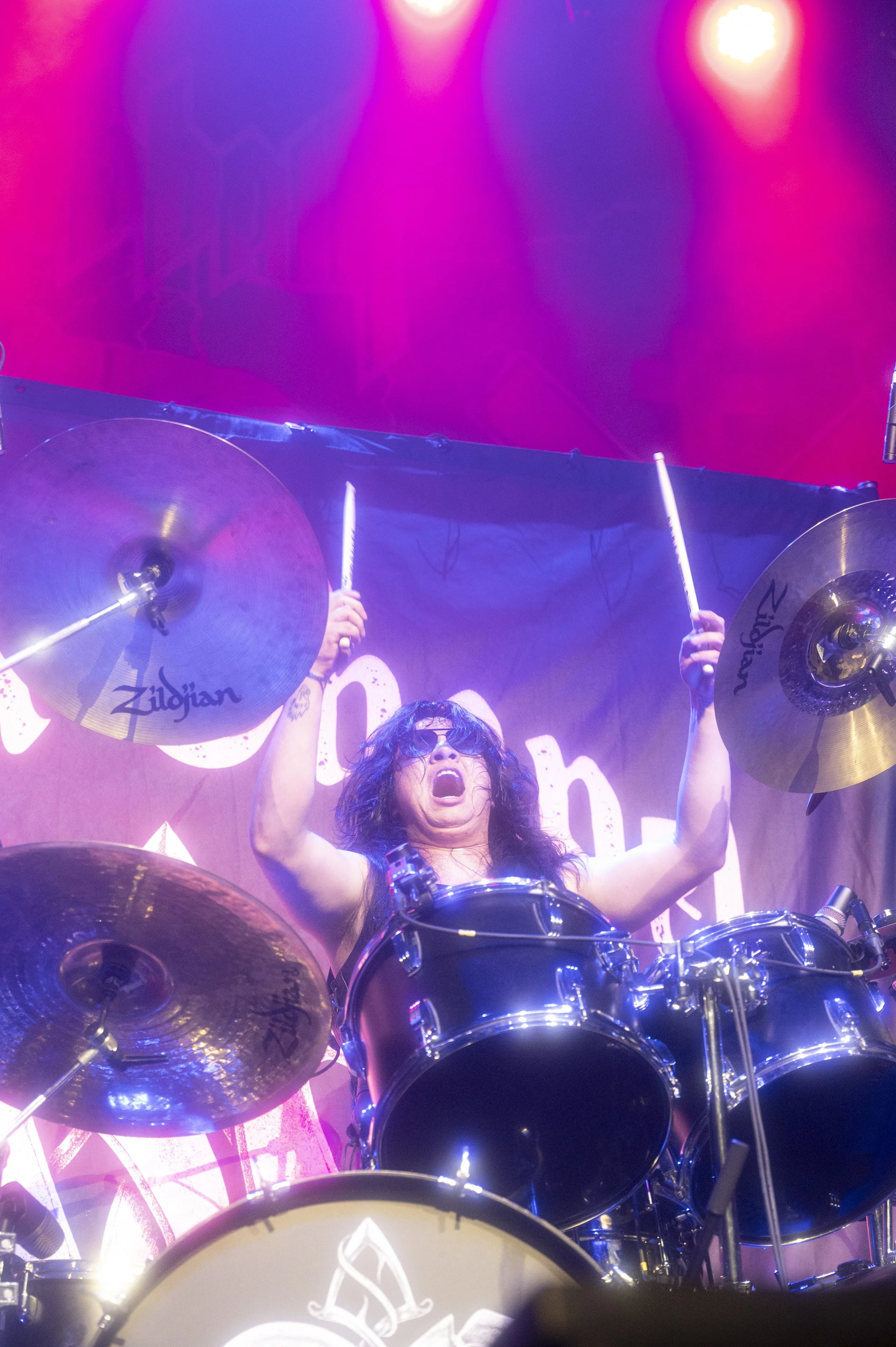 A woman with black, curly hair and sunglasses playing a drum set on stage, raising drumsticks in the air, with colorful stage lighting overhead