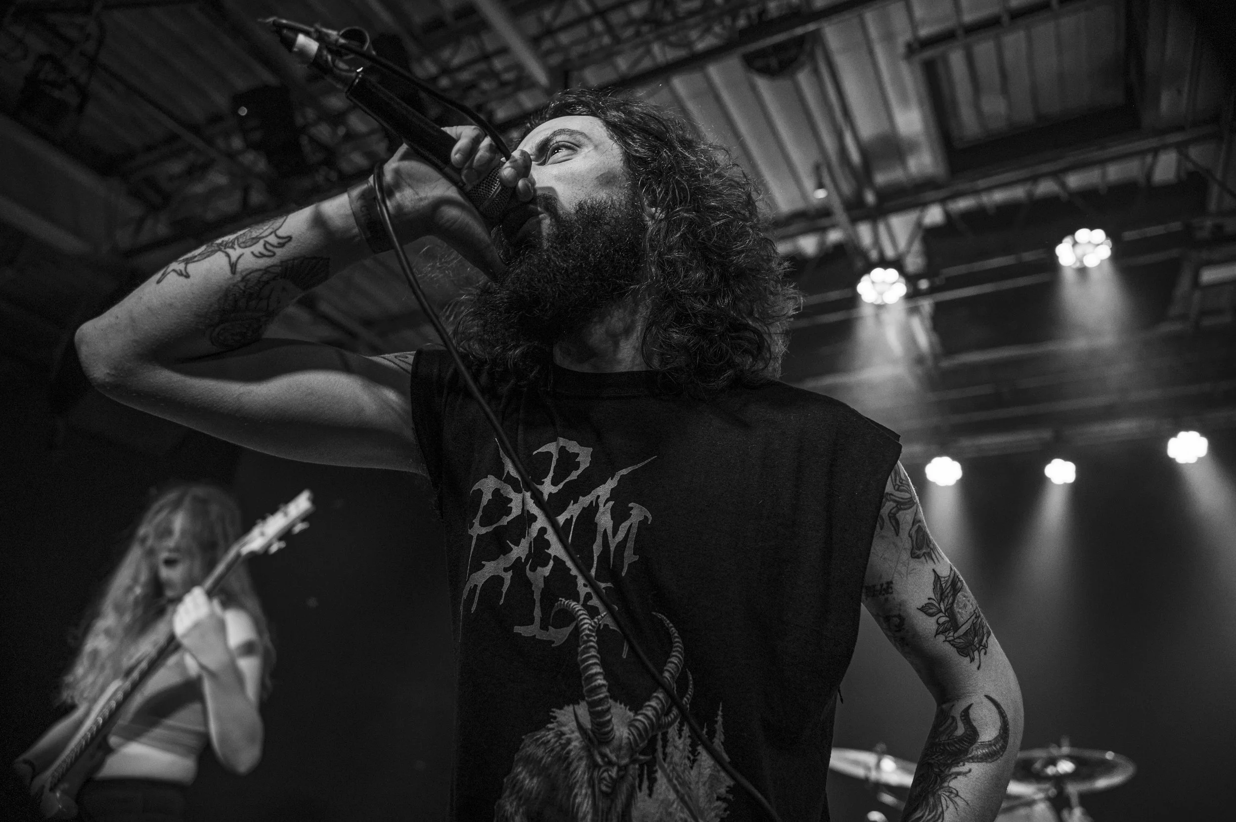 A male singer with tattoos, curly hair, and a beard performing on stage, holding a microphone close to his mouth, while a female guitarist with long hair plays in the background, in a dimly lit concert setting.