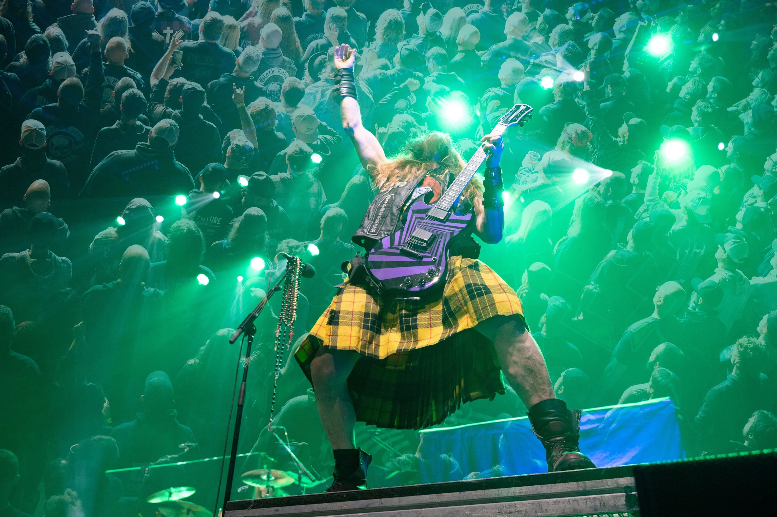 A musician with long hair wearing a kilt and leather boots playing a purple guitar on stage at a concert, with a large crowd in the background.