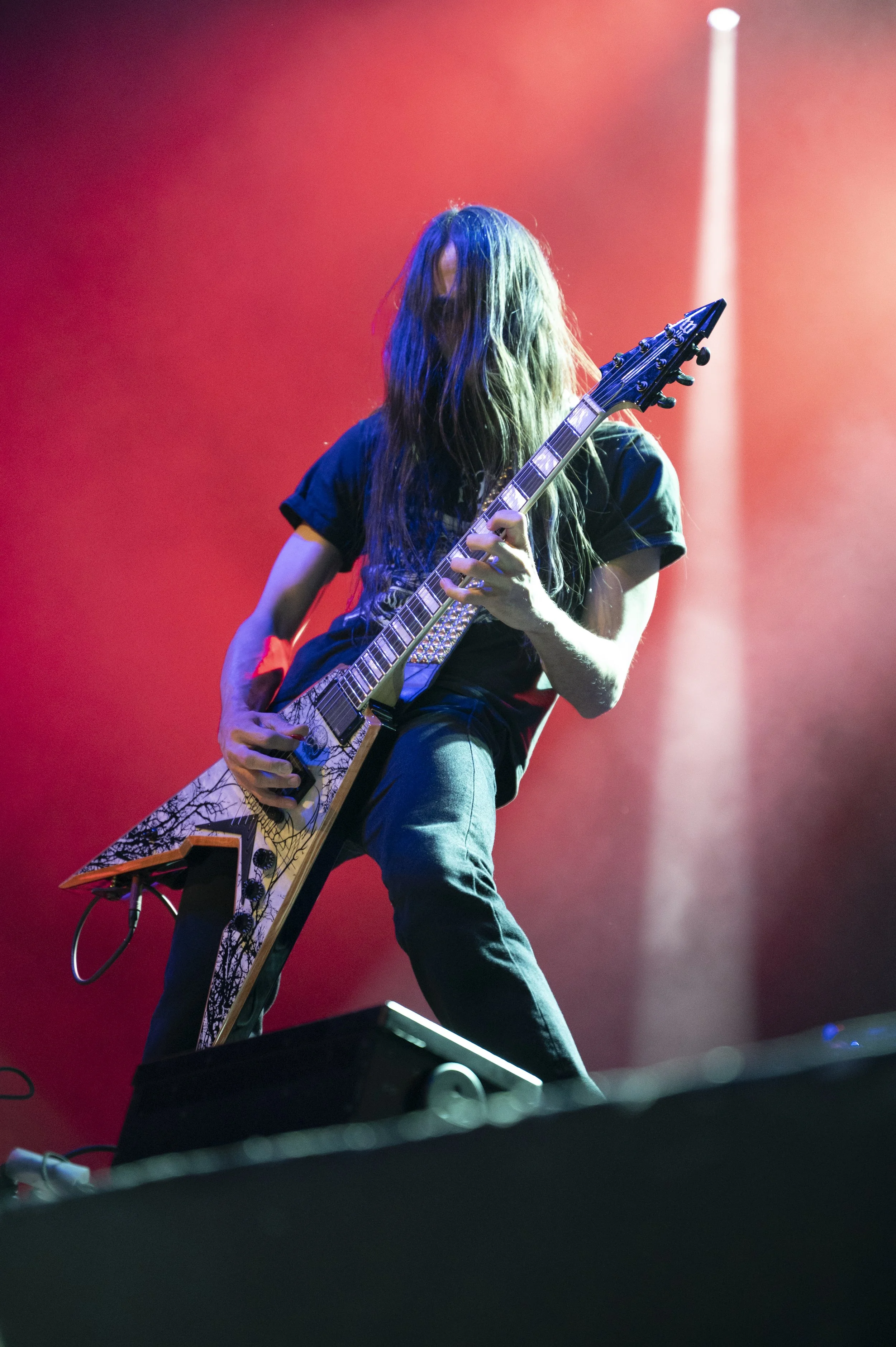 A long-haired guitarist playing an electric guitar on stage with red lighting in the background.