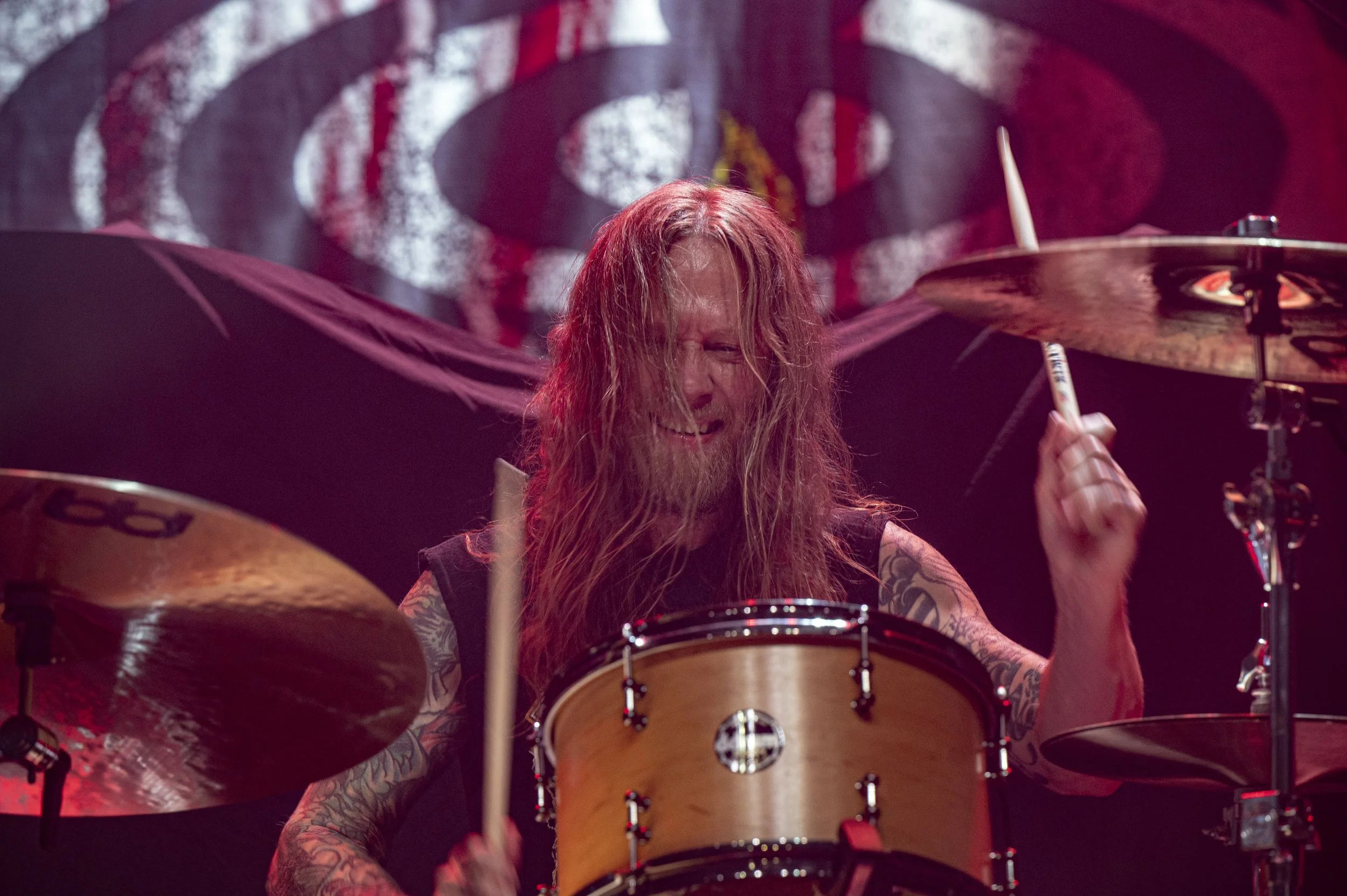 A long-haired drummer with tattoos, passionately playing a drum set on stage with a backdrop showing the Red Hot Chili Peppers logo.