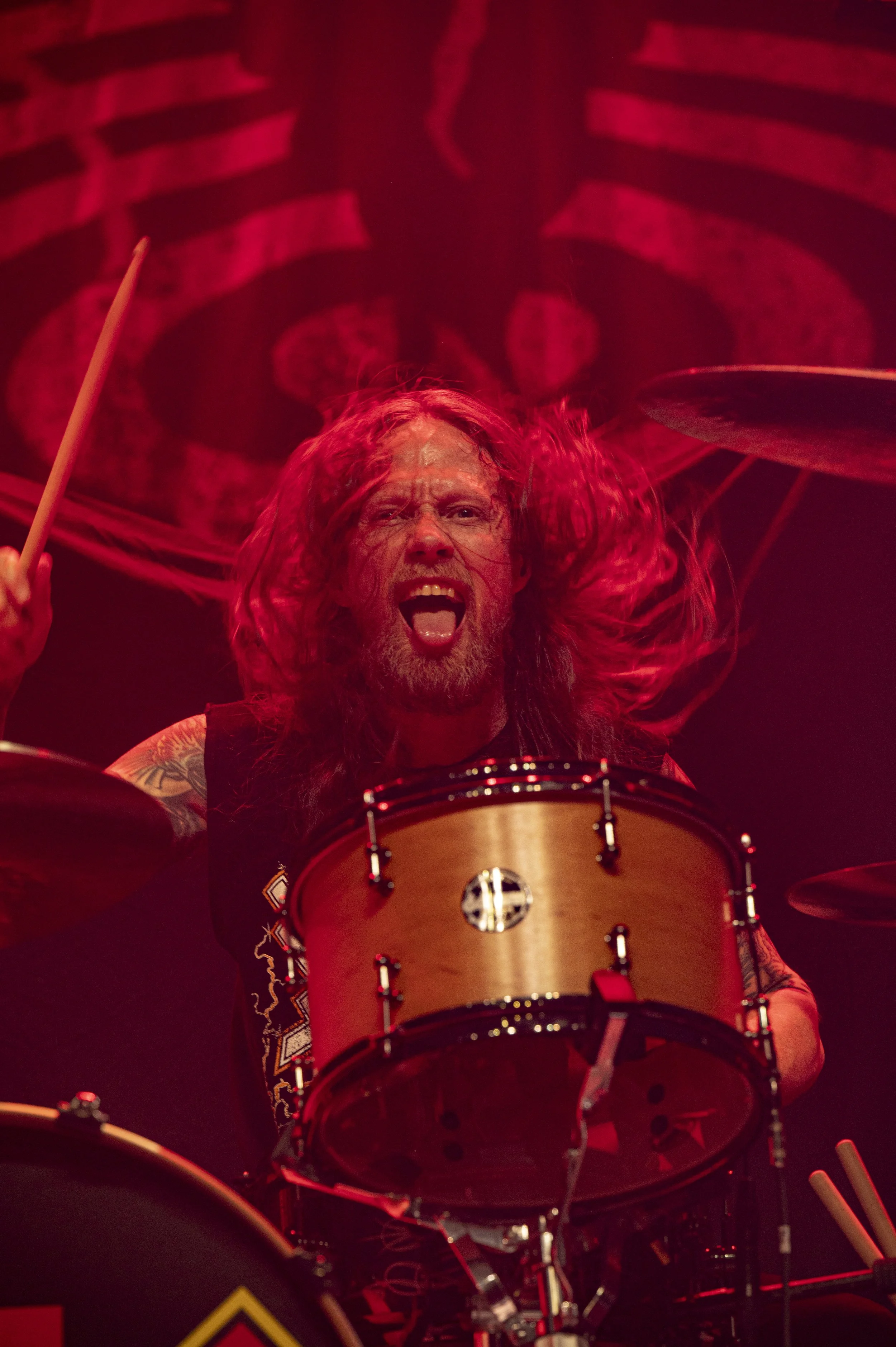 A man with long hair, a beard, and tattoos is passionately playing the drums during a live concert, with a dark background and red lighting.