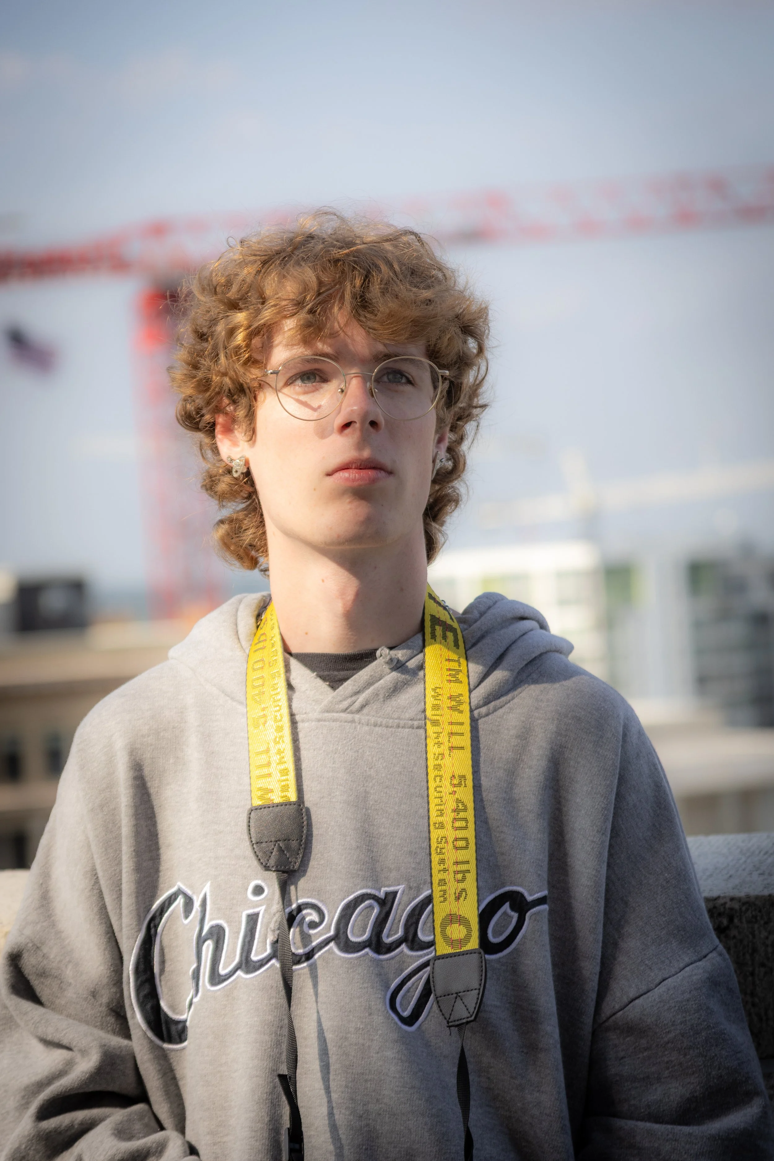 A young man with curly hair wearing glasses and a hoodie with 'Chicago' written on it, standing outdoors with a construction crane and buildings in the background.