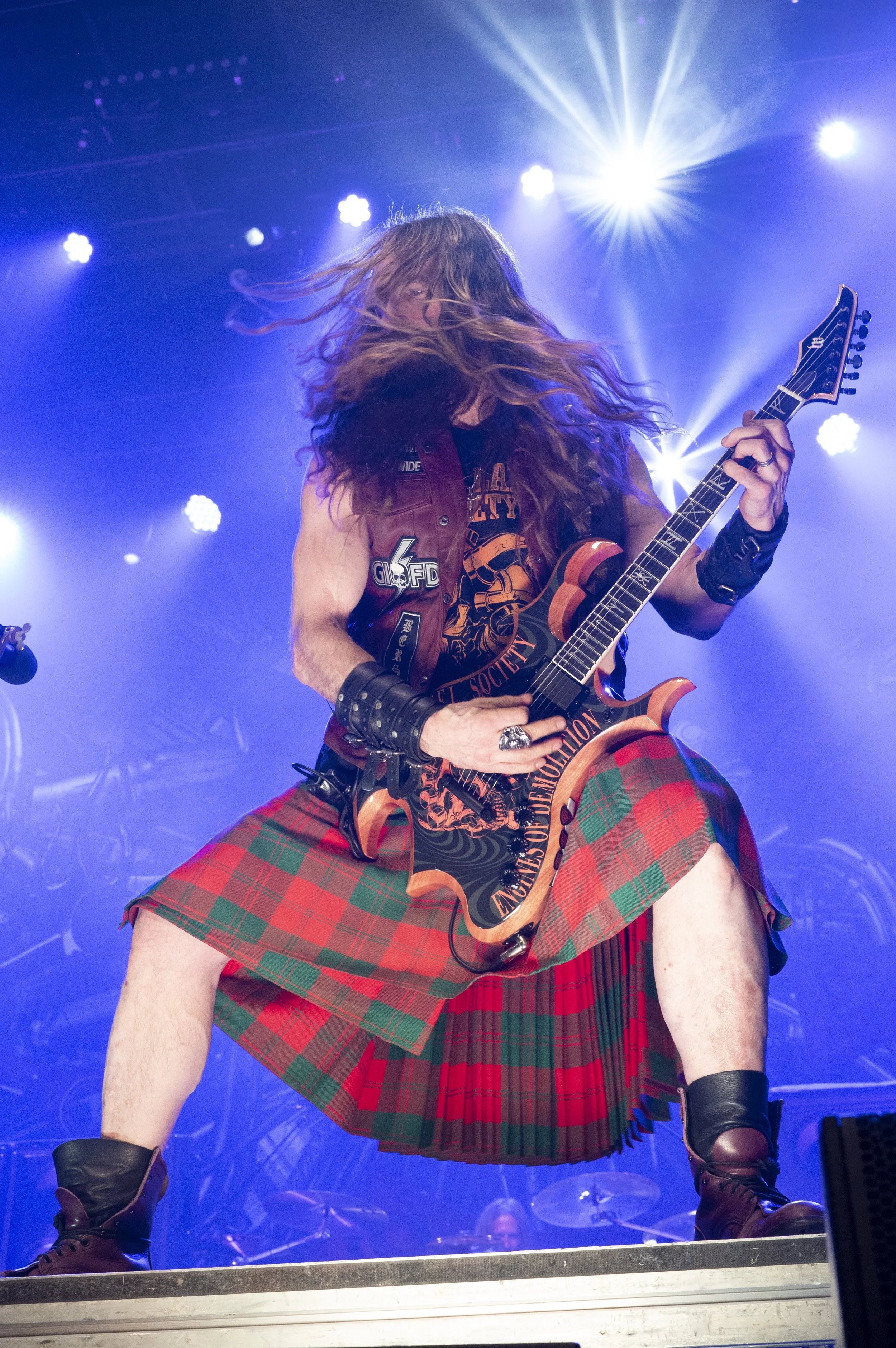 A guitarist wearing a Scottish kilt, leather wristbands, and a vest performing on stage with lighting effects and a drum set in the background.