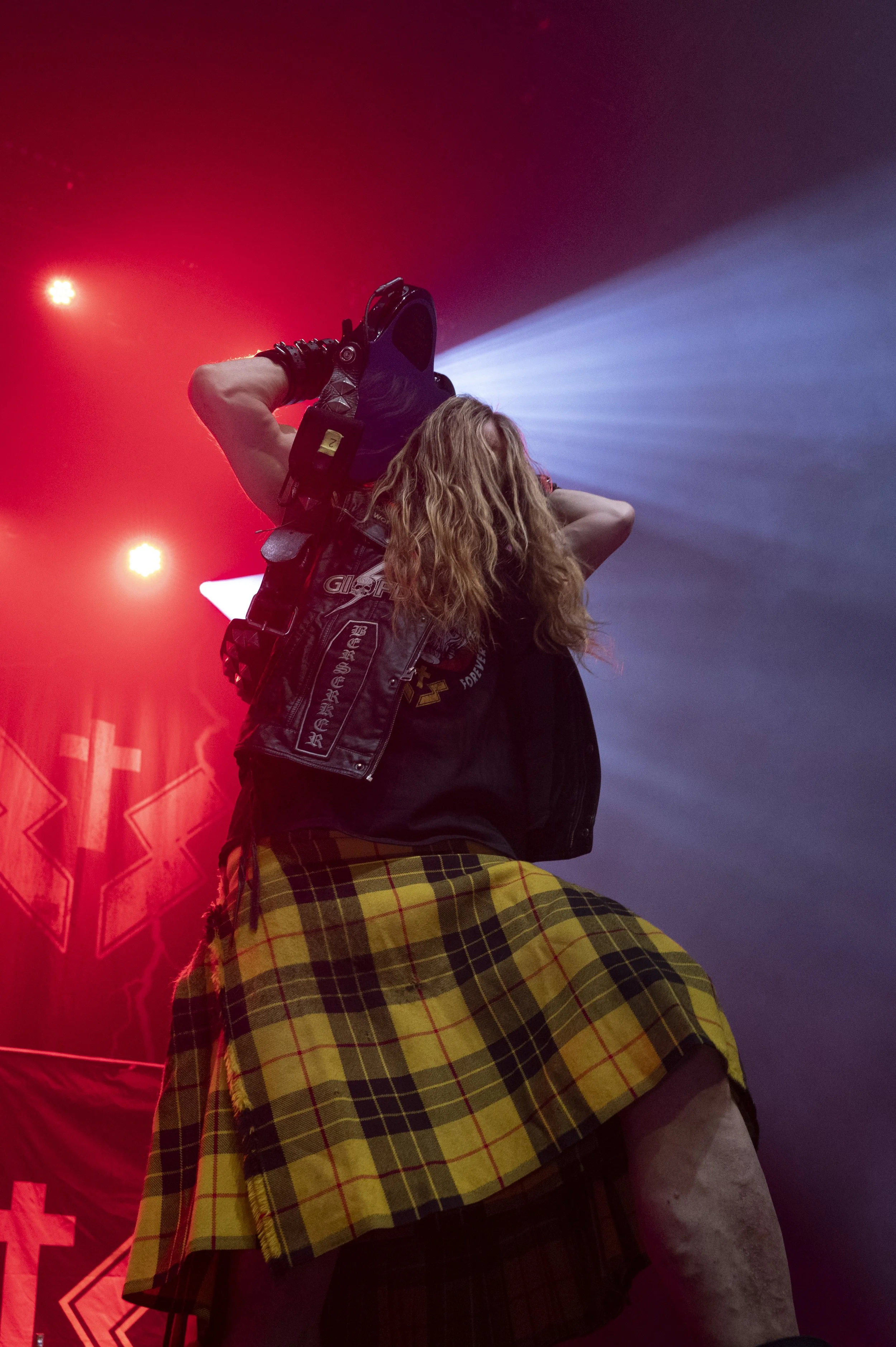 A person with long blonde hair taking a photograph with a camera on a concert stage. They are wearing a black vest with patches and a yellow plaid kilt, with stage lights and a backdrop behind them.