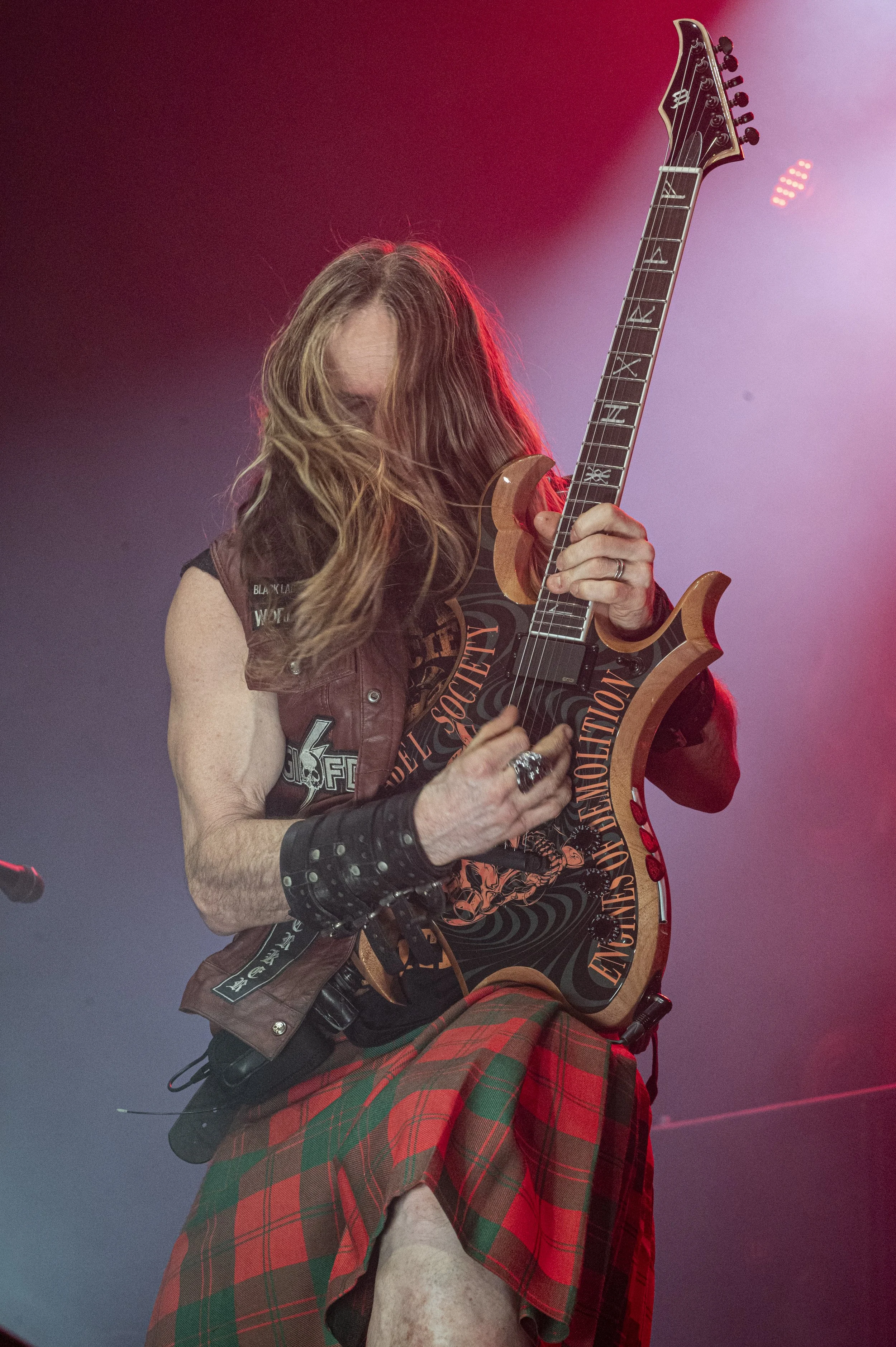 A long-haired musician dressed in a kilt and a leather vest playing an electric guitar on stage.