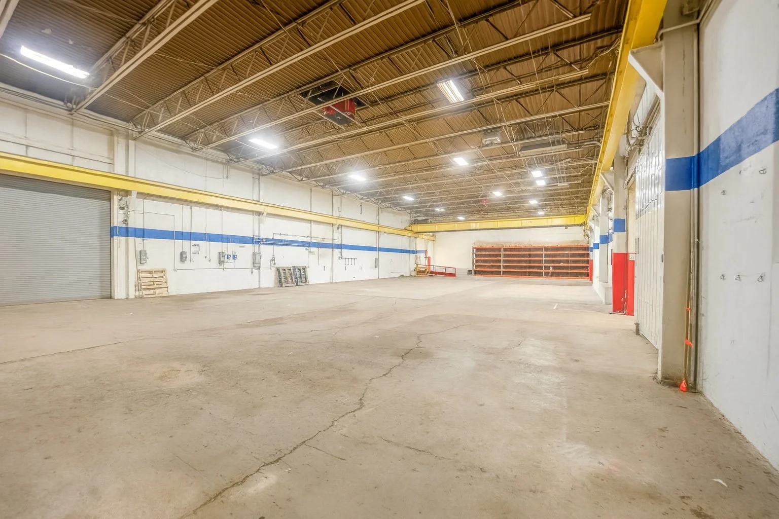 Empty industrial warehouse with concrete floor, yellow and blue safety barriers, and overhead lights.