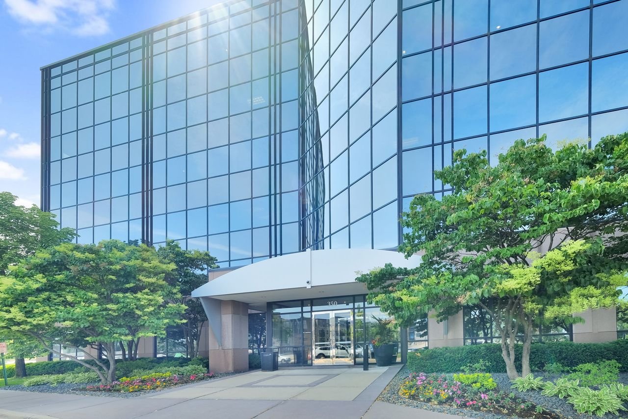 Modern office building with extensive glass windows and a landscaped entryway with trees and flowers.