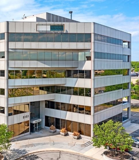 Modern multi-story office building with reflective glass windows, located at 5151, surrounded by trees and parking lot.
