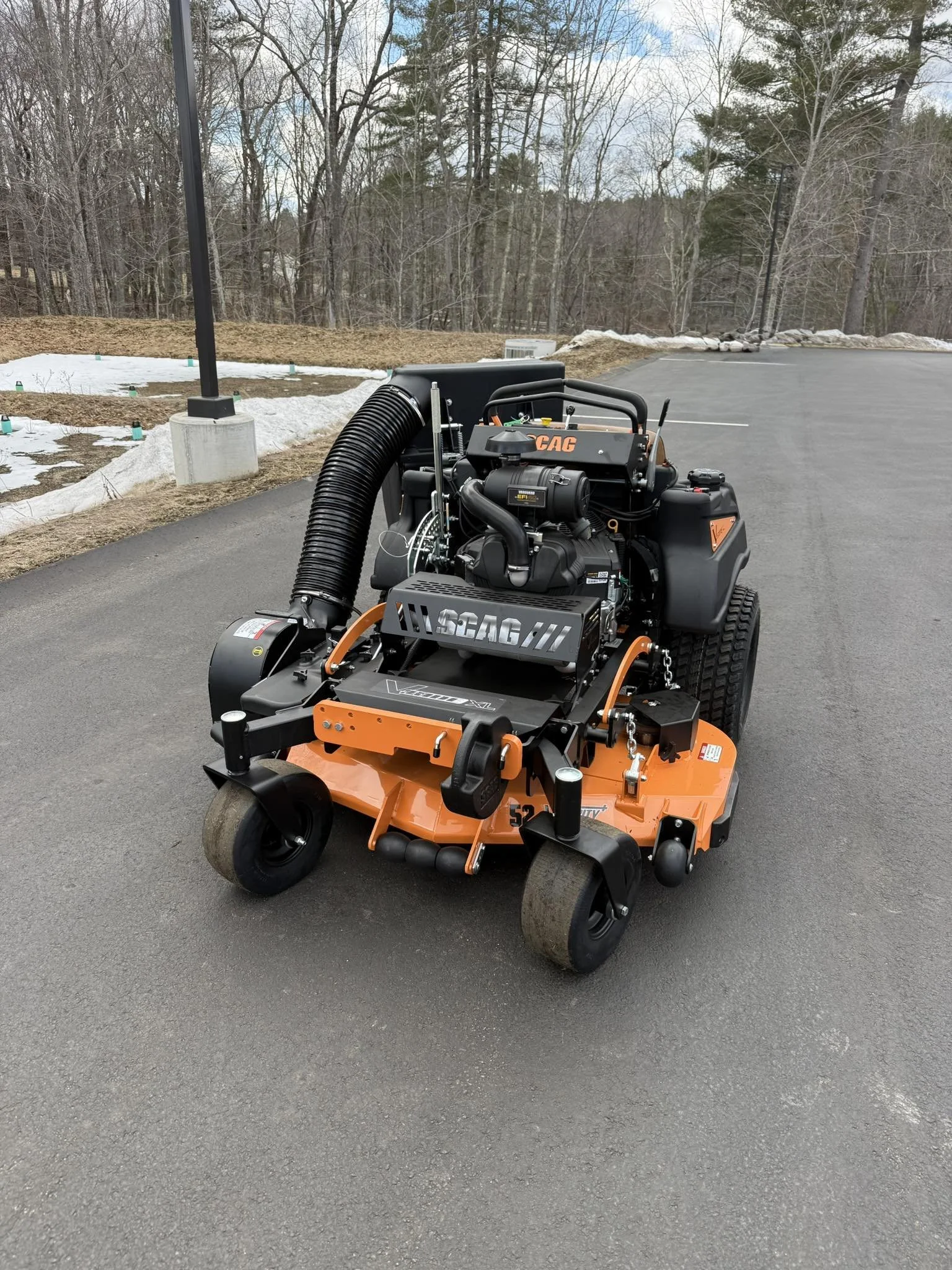 A large orange and black commercial lawn mower parked on an asphalt surface with a snowy background and trees in the distance.