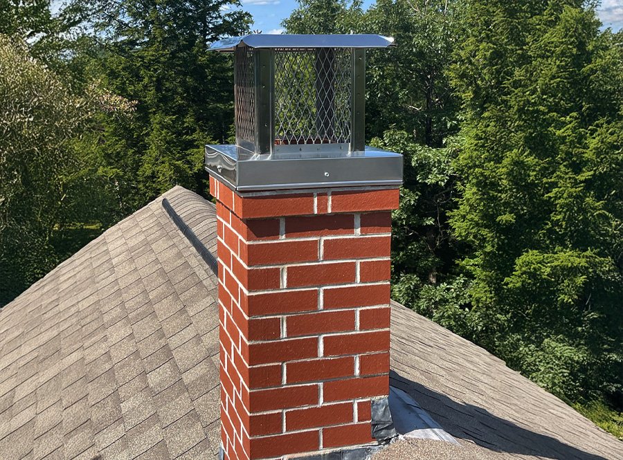 Photo of a house roof with a brick chimney that has a metal chimney cap on top, and a surrounding lush green forest in the background.