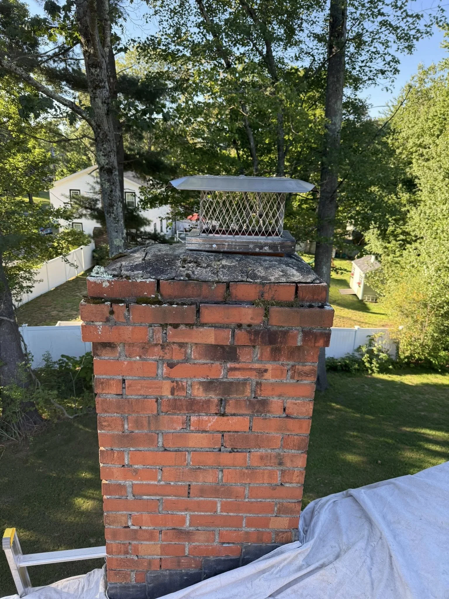 A brick chimney with a metal cap atop a house roof, surrounded by green trees and a white fence in the background.