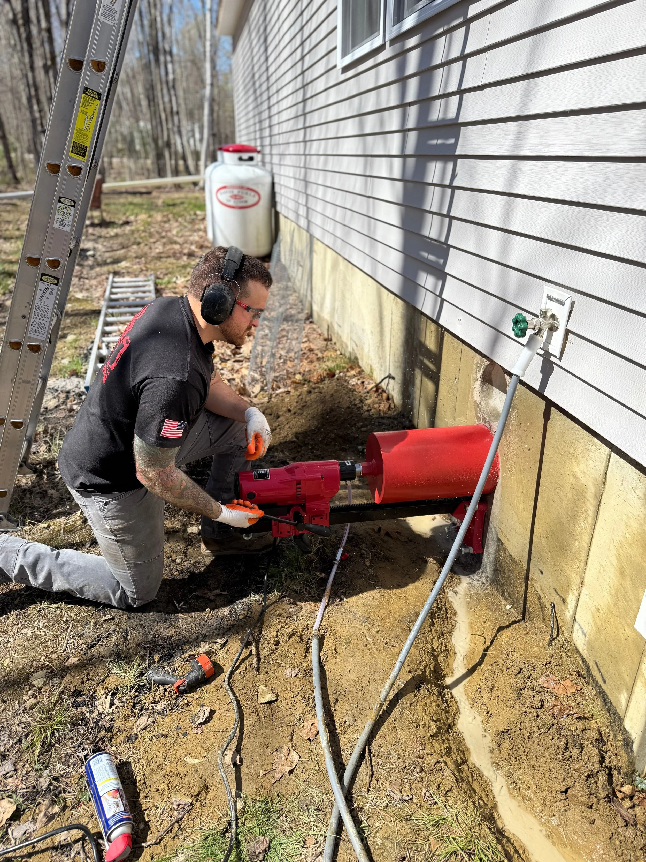 A man kneeling outdoors next to a house wall, wearing ear protection and gloves, using a red electric jack hammer to work on the foundation near a water spigot with exposed pipes, surrounded by tools and equipment on the ground.