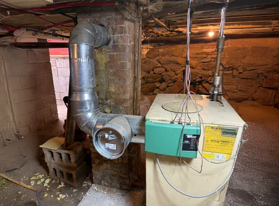 An HVAC furnace system with a metal vent pipe, part of the vent pipe is wrapped around a brick and concrete pillar in a basement. There are wires hanging from above, and the furnace has labels and control boxes attached.