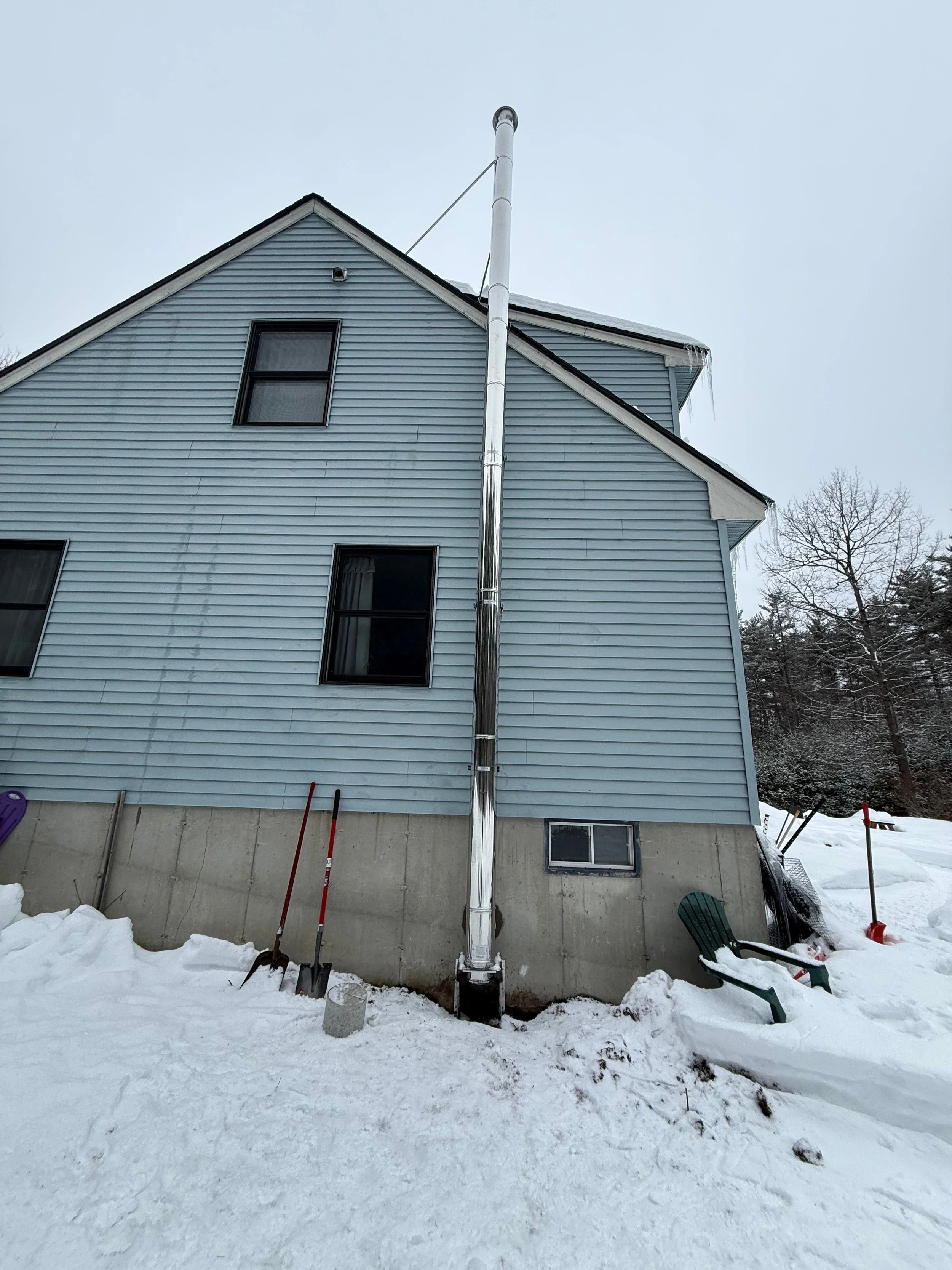 Side view of a blue house with two windows, snow on the ground, and a tall metal chimney pipe extending from the foundation to above the roof, with icicles hanging from the eaves.