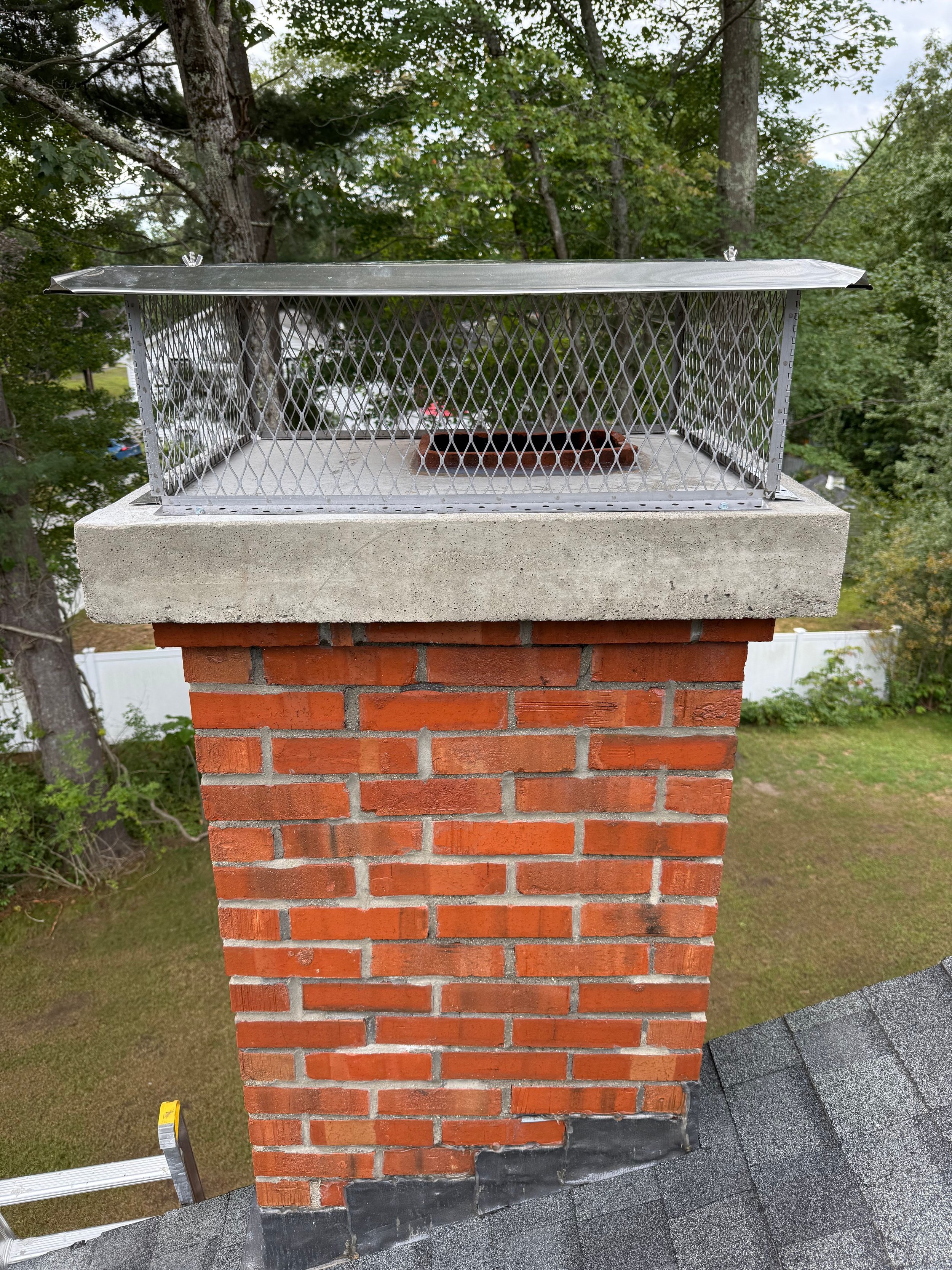 A brick chimney with a metal chimney cap on top, situated on a shingled roof surrounded by trees.