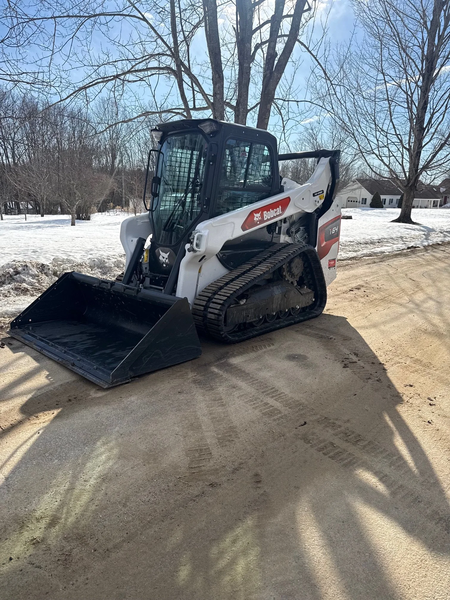 A Bobcat T54 compact track loader parked on a dirt path, with snow and trees in the background on a clear day.