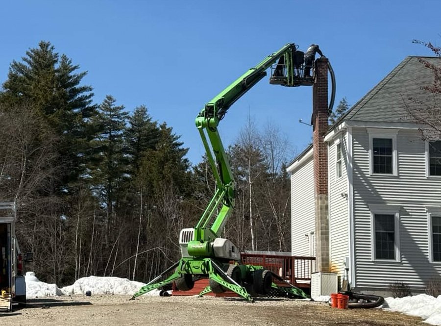 A green boom lift extended to reach the top of a brick chimney on a two-story white house during daytime.
