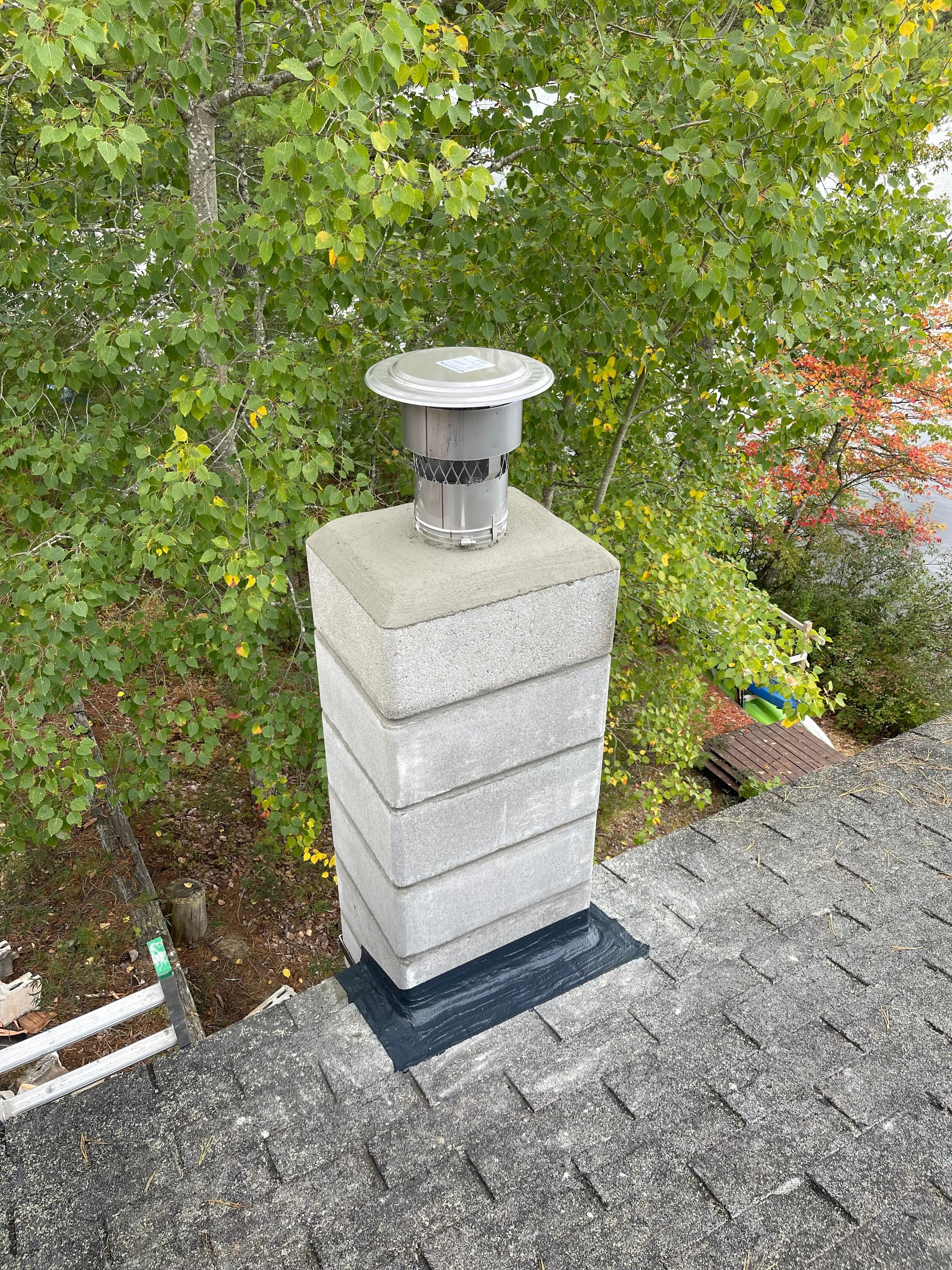 A metal chimney cap on top of a brick chimney on a shingled roof, with green leafy trees in the background.